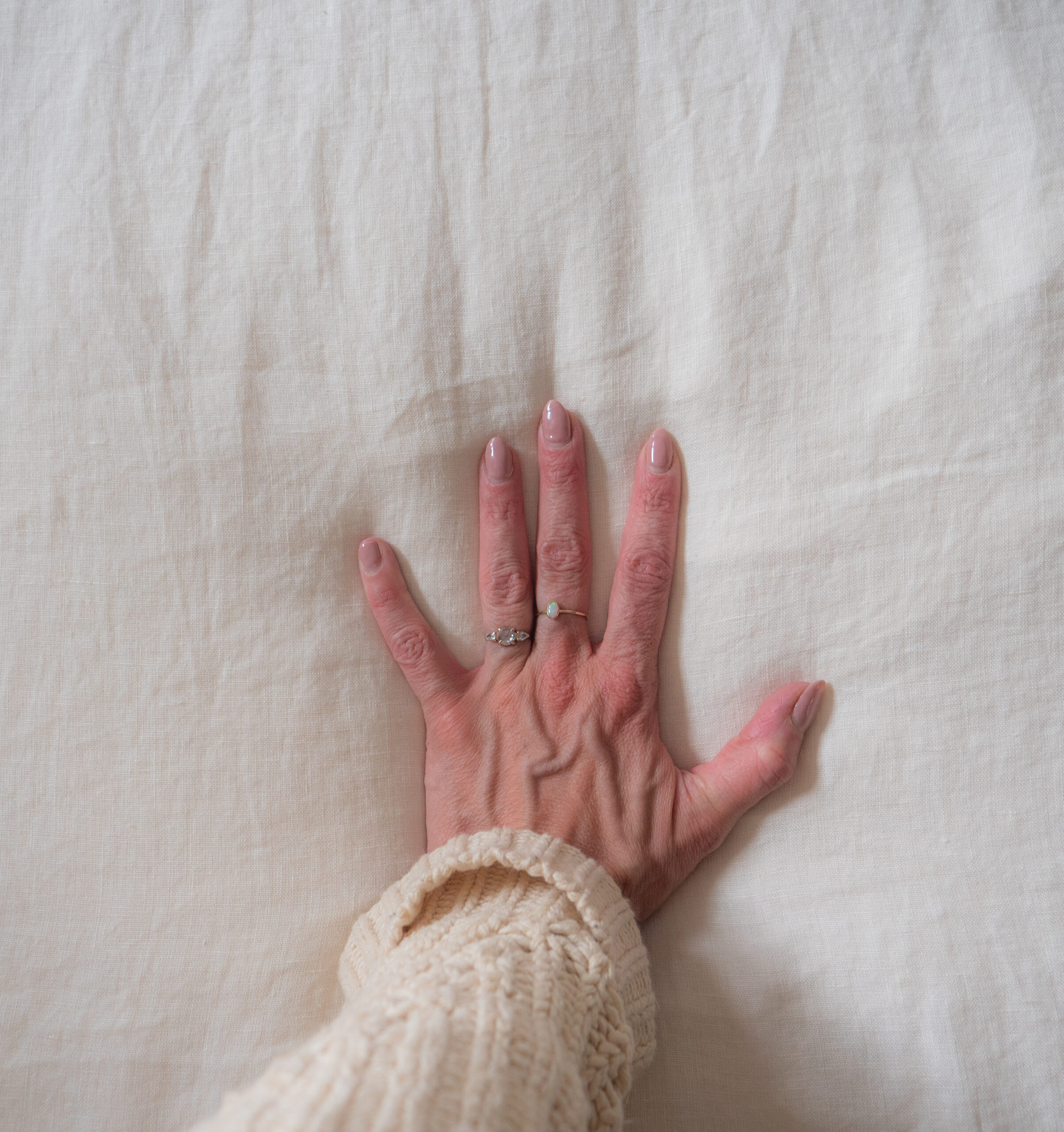 A hand wearing two rings rests palm-down on a cream-colored textured fabric background.