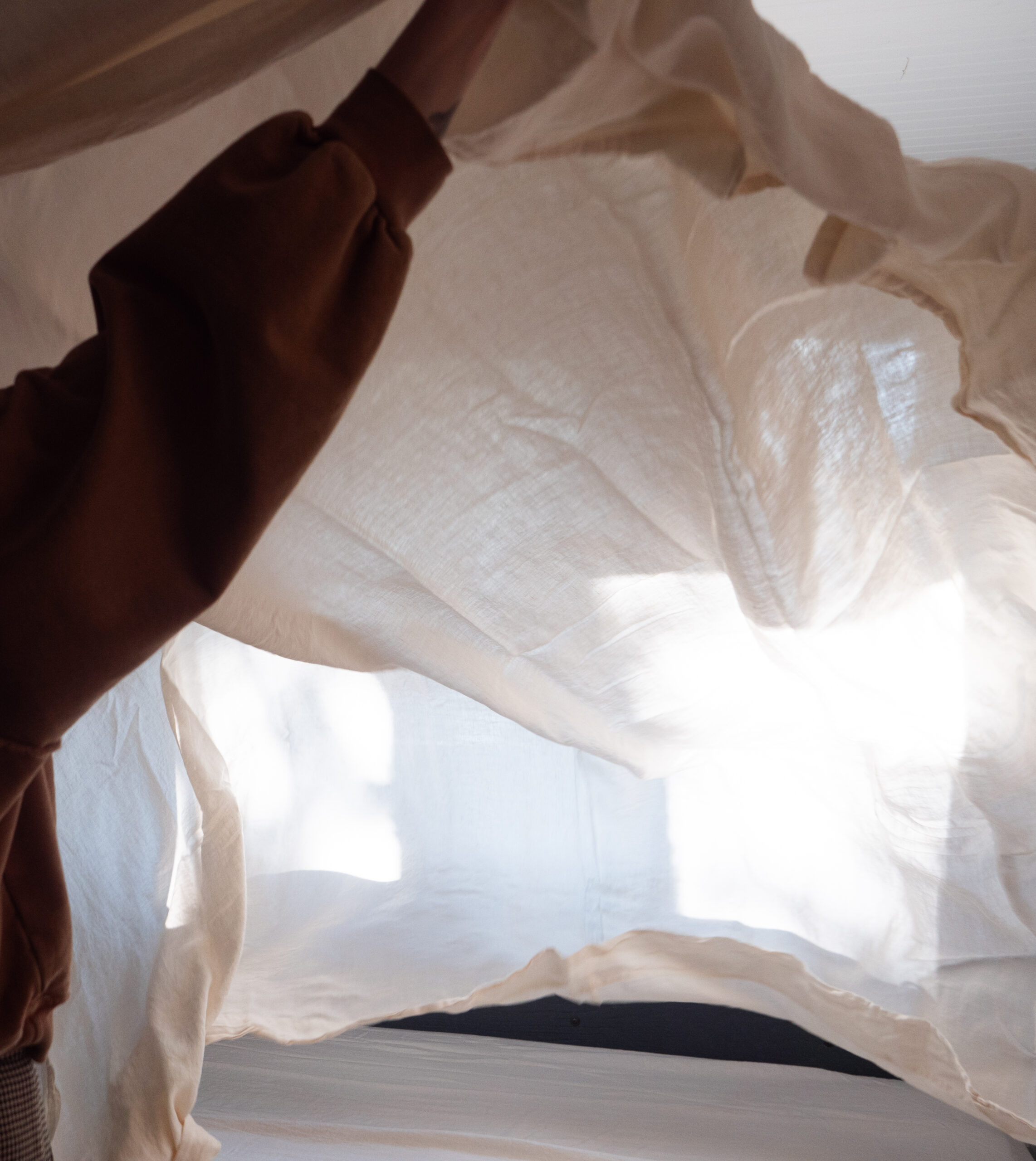 A person in a brown sleeve holds up a light, sheer fabric near a window with sunlight streaming through.