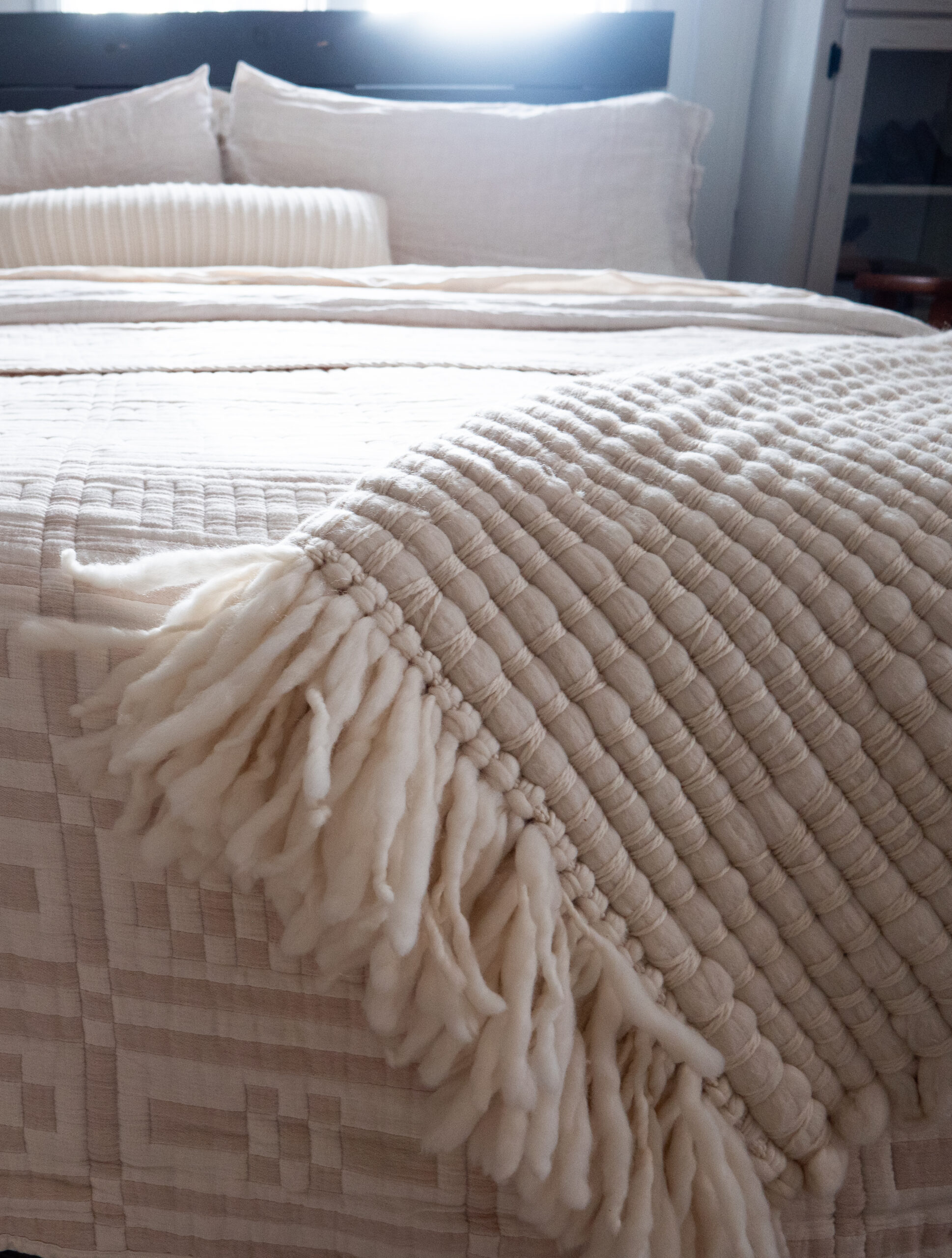 A neatly made bed with cream-colored pillows, a textured quilt, and a thick, fringed knit throw draped at the foot.