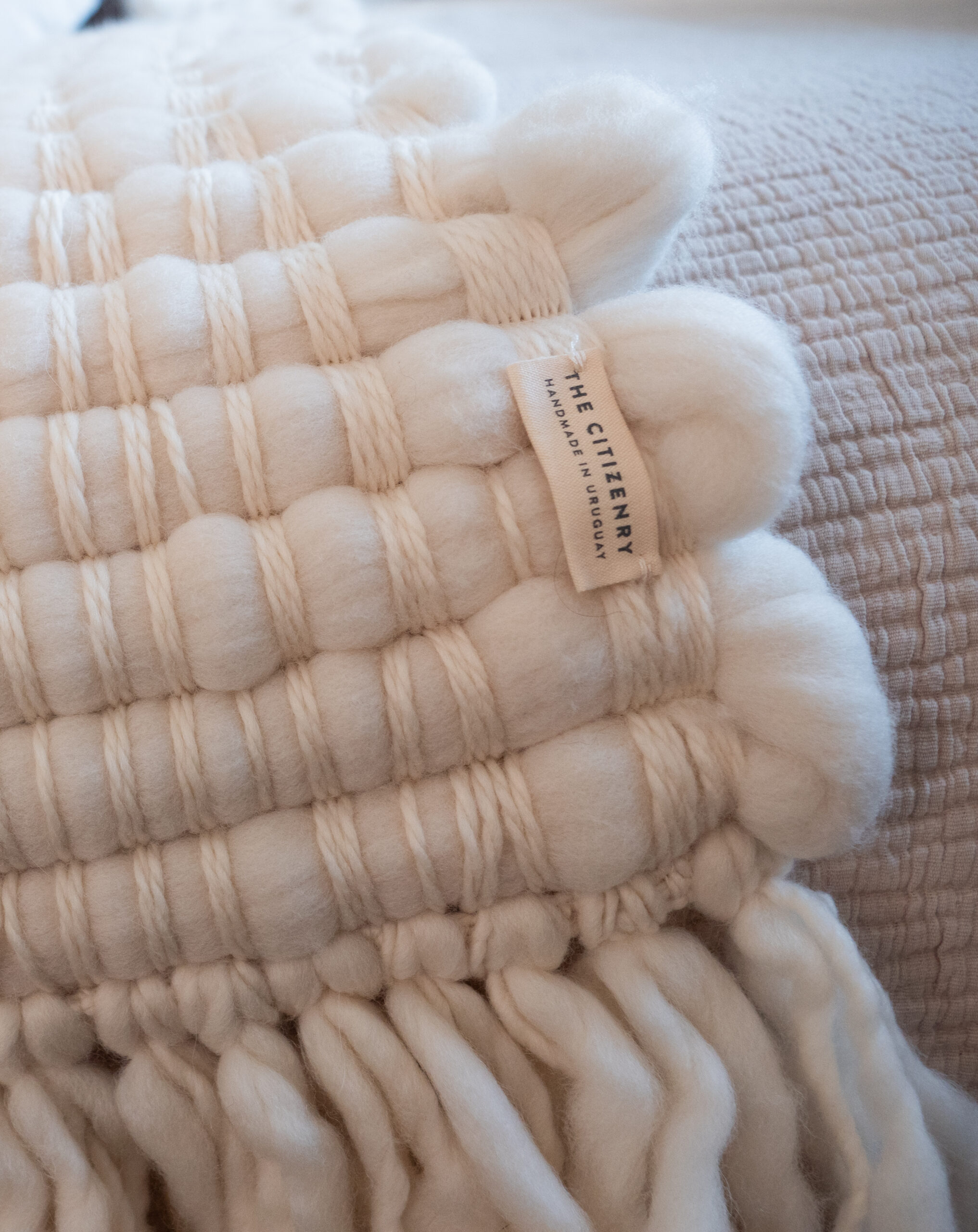 A close-up of a textured cream-colored woven throw blanket with thick yarn, a fringed edge, and a visible label reading "THE CITIZENRY.