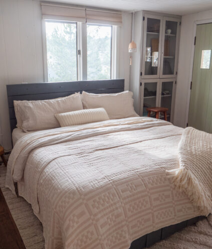 A neatly made bed with cream-colored bedding and pillows sits in a bright room with two windows, a glass cabinet, and a green door.