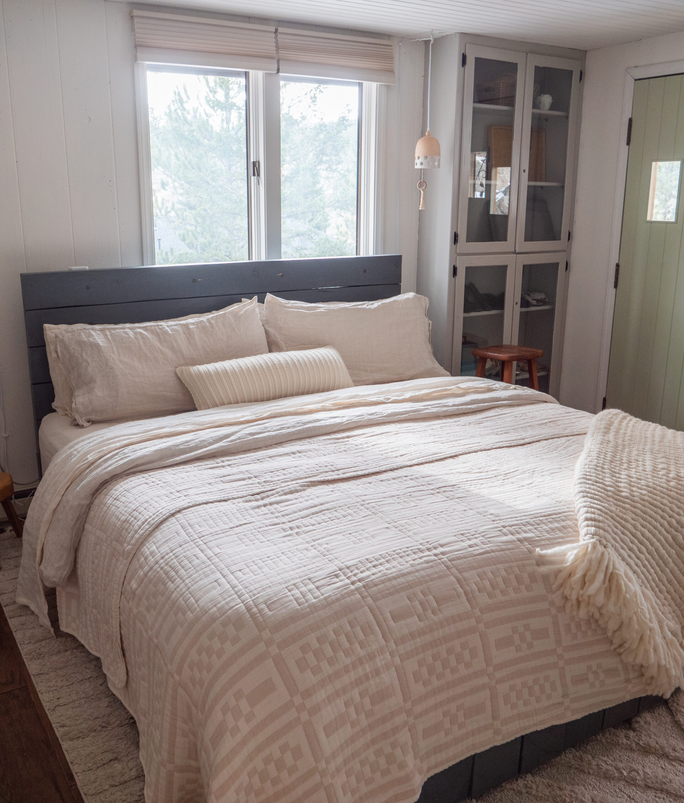A neatly made bed with cream-colored bedding and pillows sits in a bright room with two windows, a glass cabinet, and a green door.
