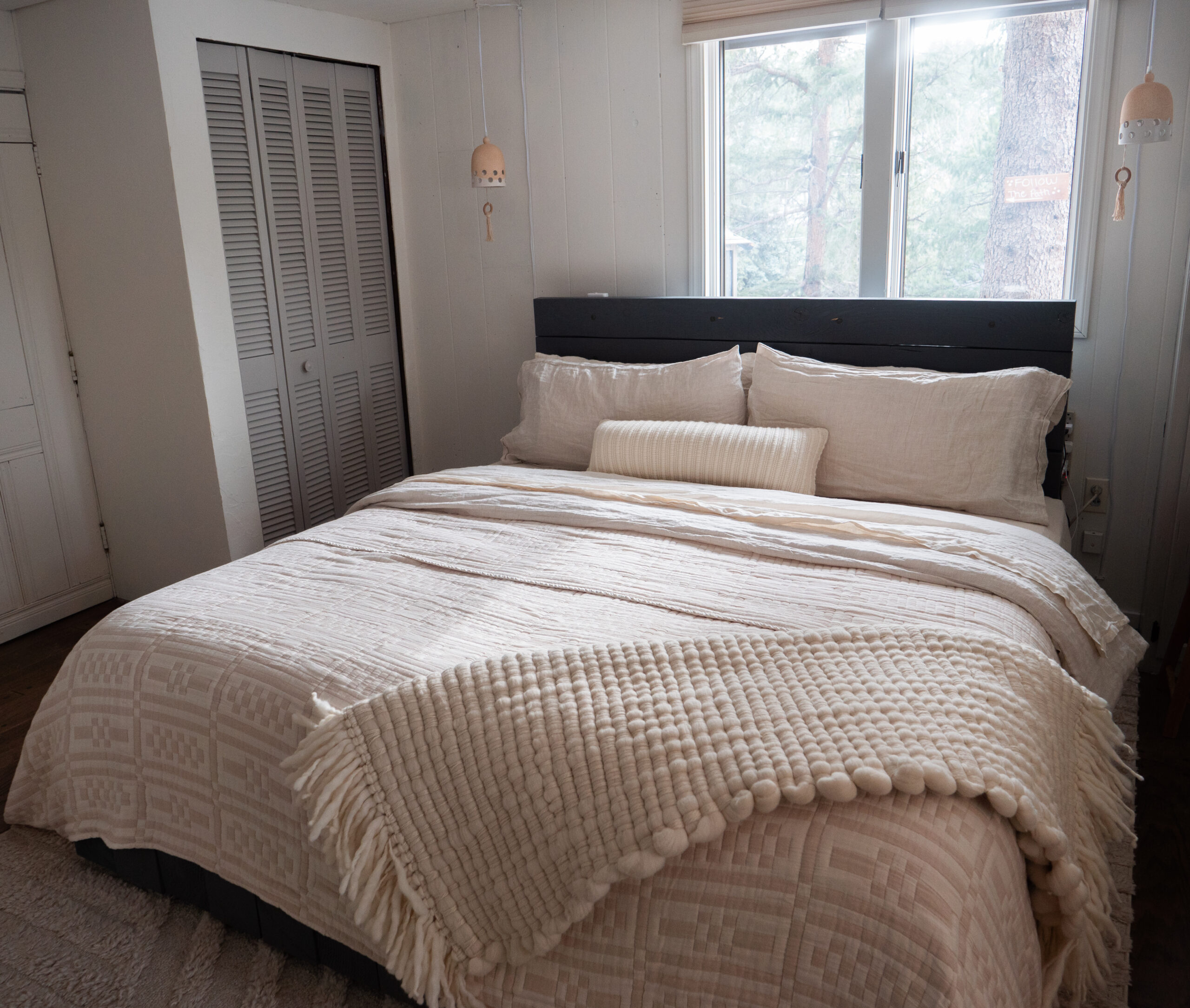 A neatly made bed with textured cream bedding and a long pillow, positioned near windows and a closed closet in a simply decorated bedroom.