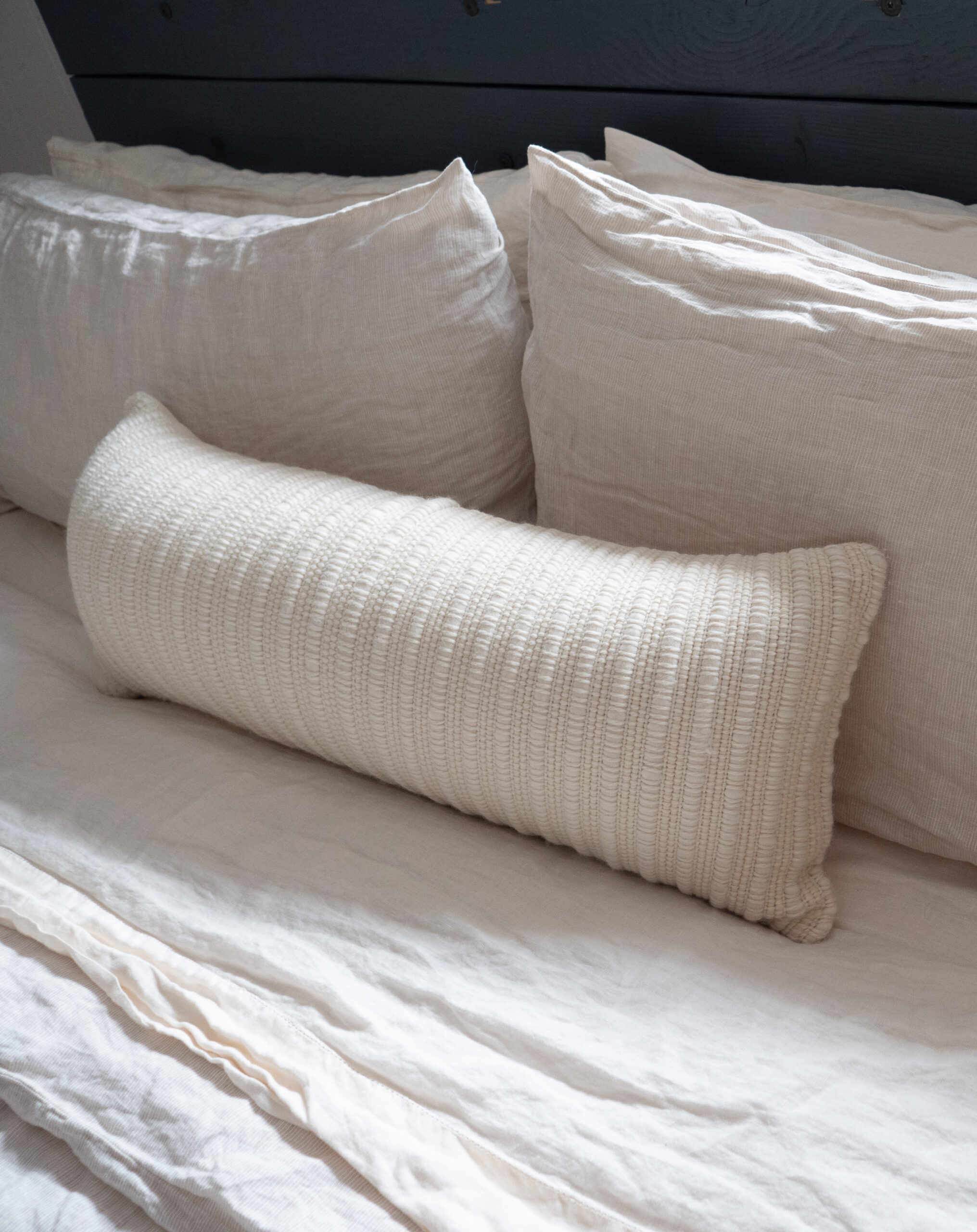 A long, ribbed white pillow rests on a bed with three large, white linen pillows and a white duvet.