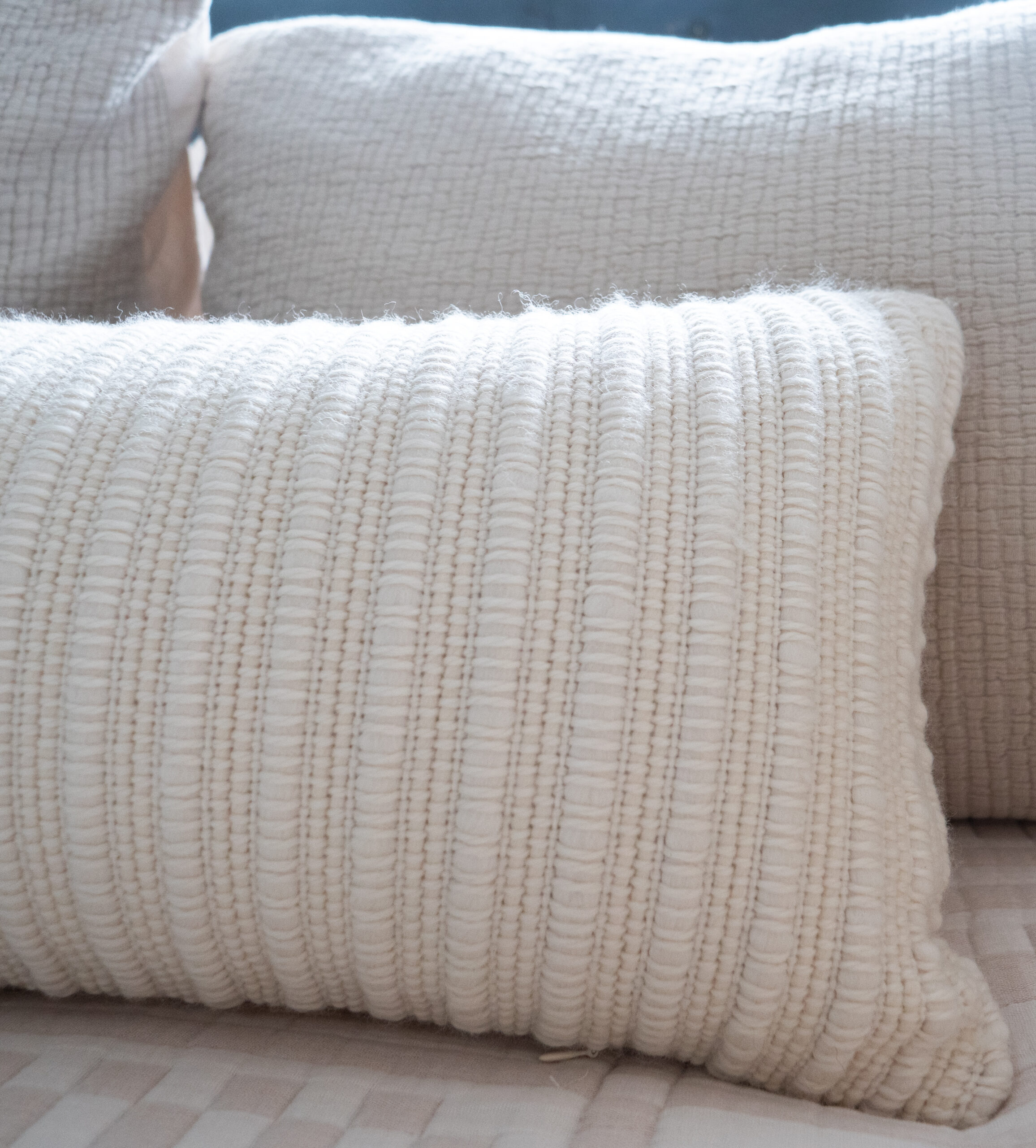 Close-up of a textured, cream-colored woven pillow in front of similarly colored, quilted pillows on a bed.