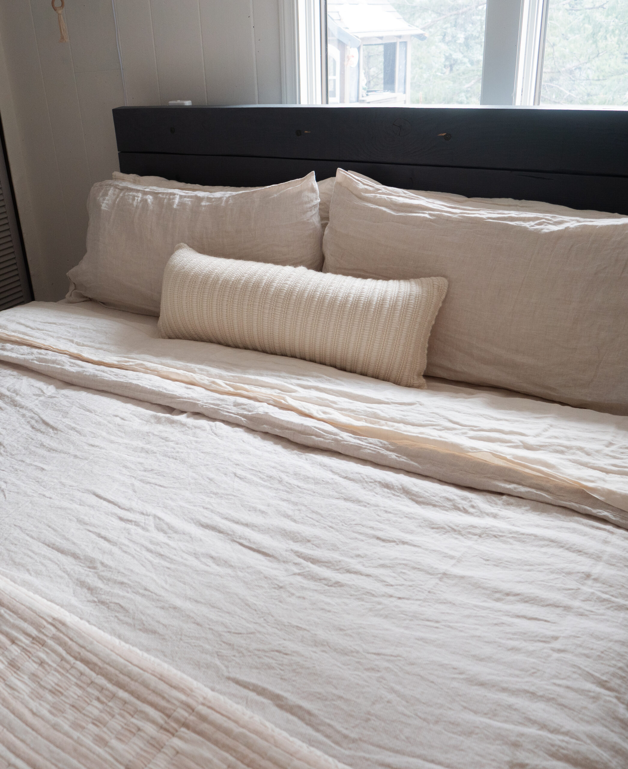 A neatly made bed with cream-colored linens, two large pillows, and a small cylindrical pillow in front, positioned under a window with white curtains.