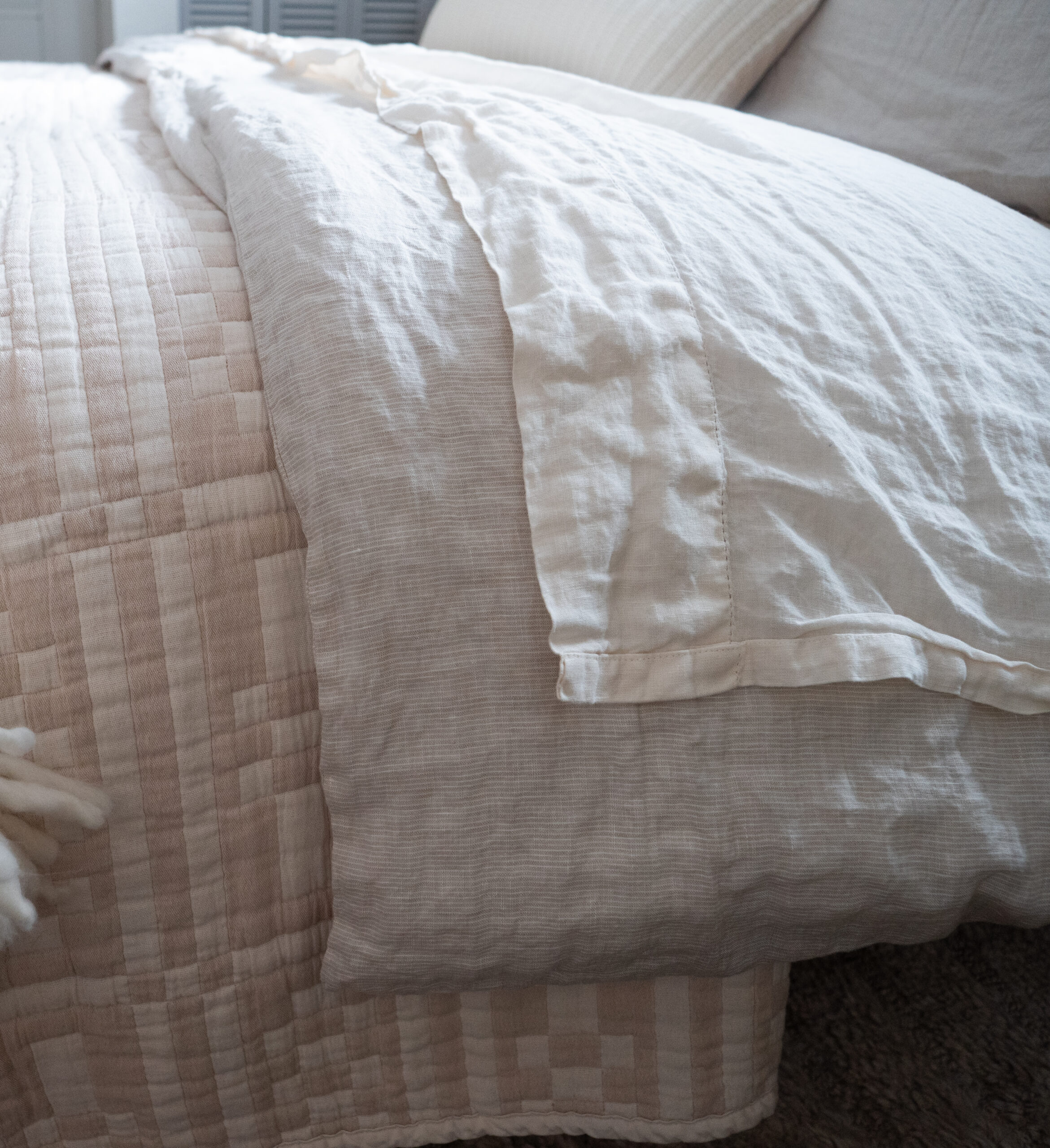 A neatly made bed with a beige quilted blanket and a white linen top sheet, positioned in a softly lit bedroom.
