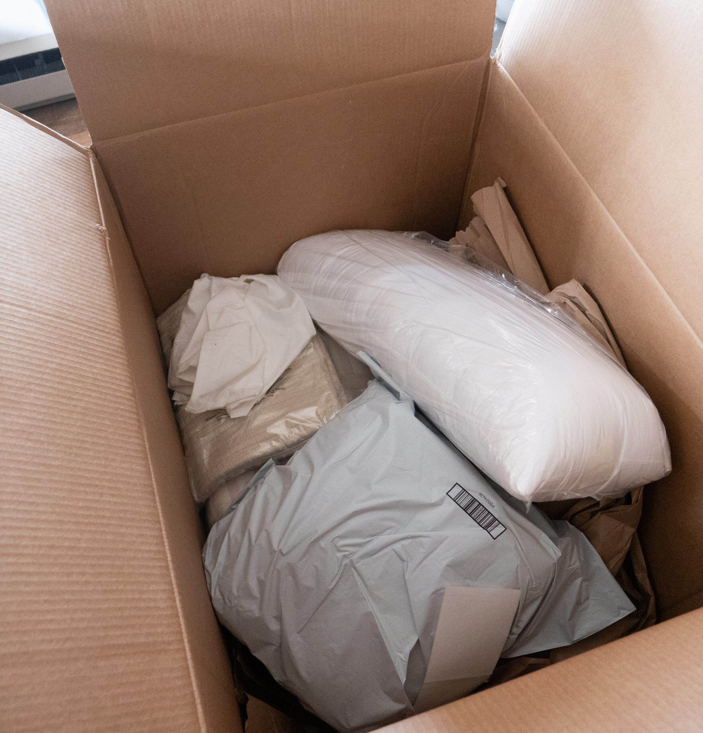 An open cardboard box containing a wrapped pillow, packaging paper, and plastic-wrapped items.