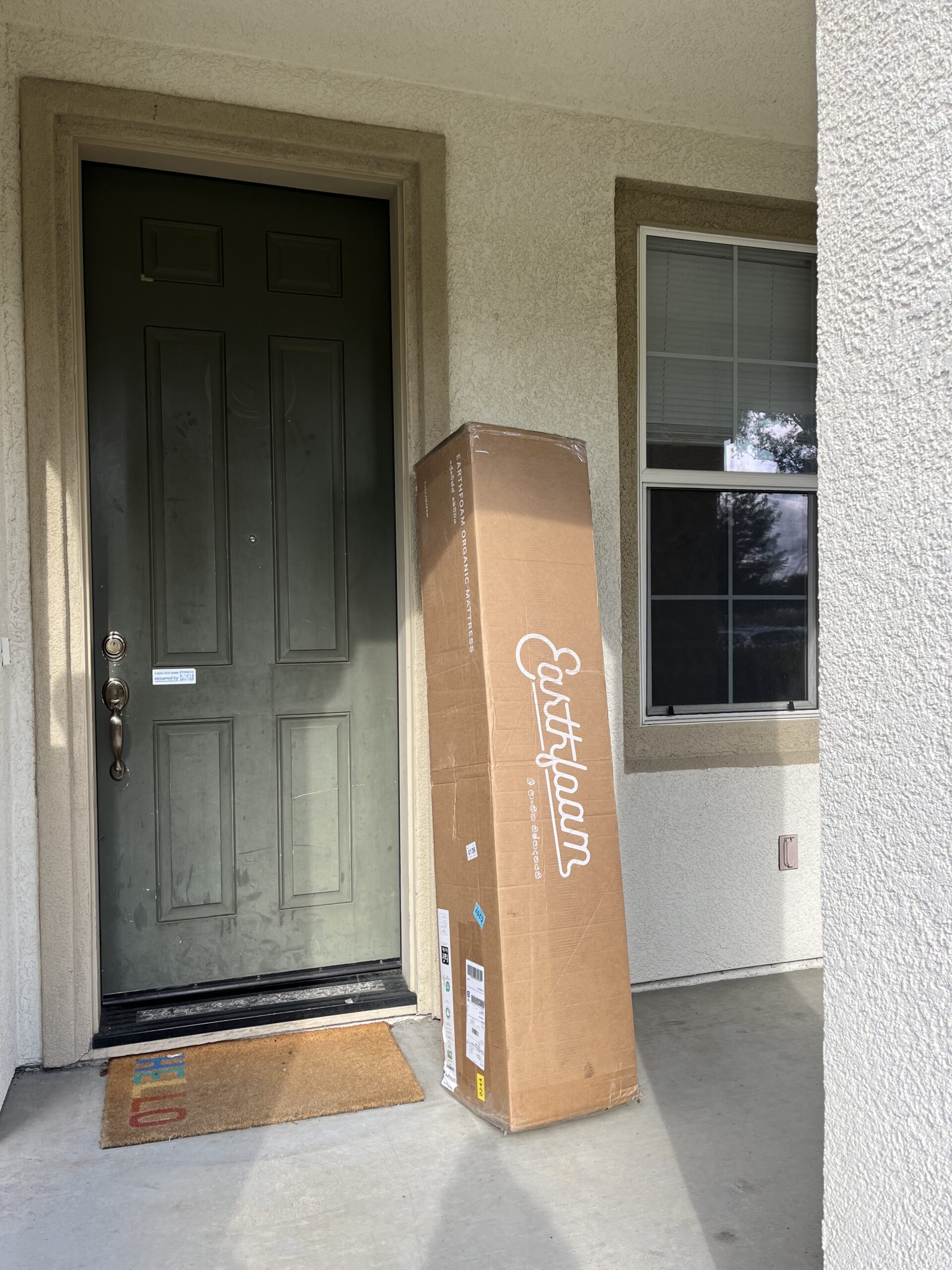 A tall cardboard box labeled "Eastbeam" resting against a closed dark green door on a front porch with a welcome mat and window nearby.