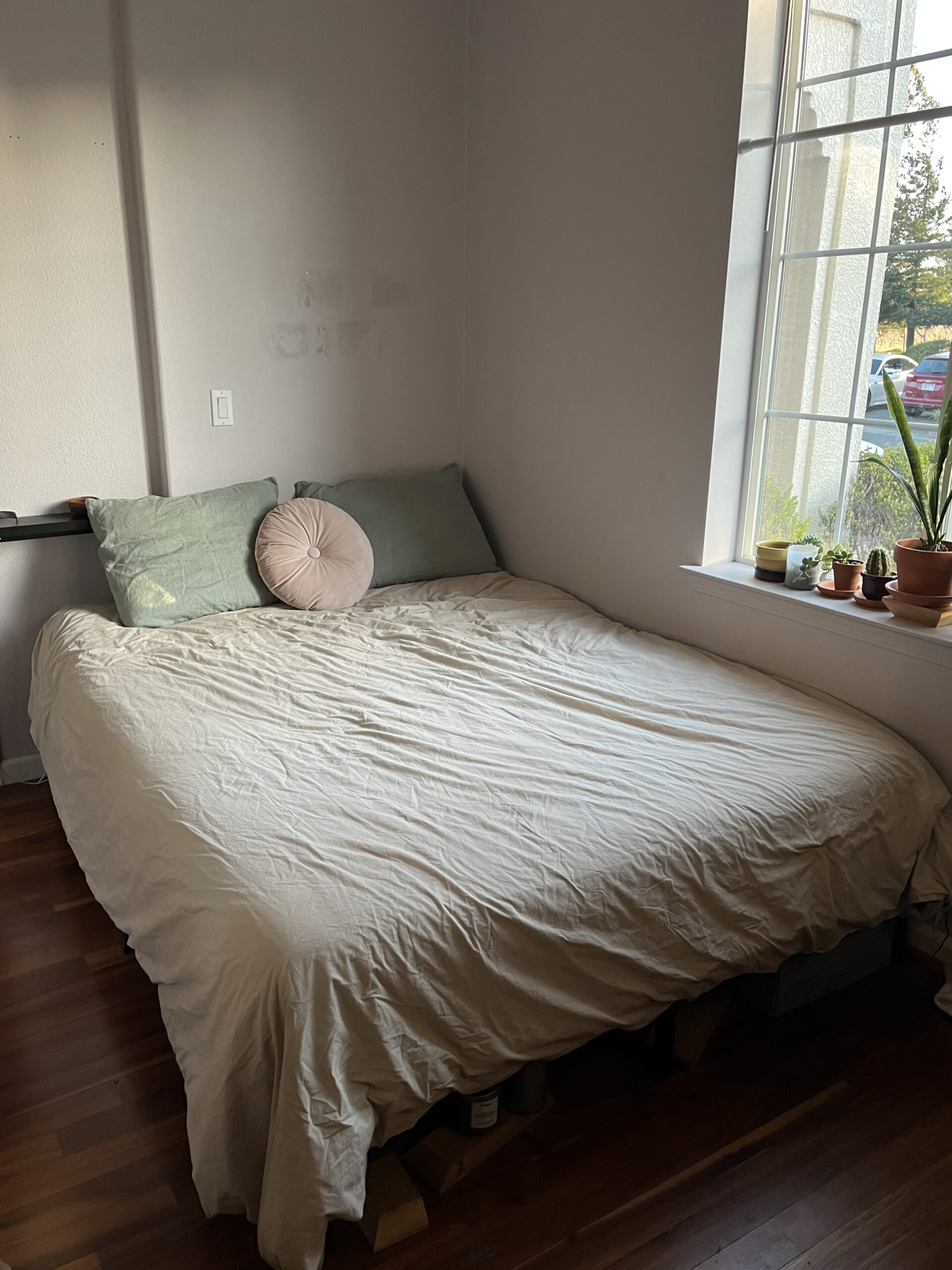 A neatly made bed with light bedding, two pillows, and a round cushion is in a corner by a large window with several small potted plants on the sill.