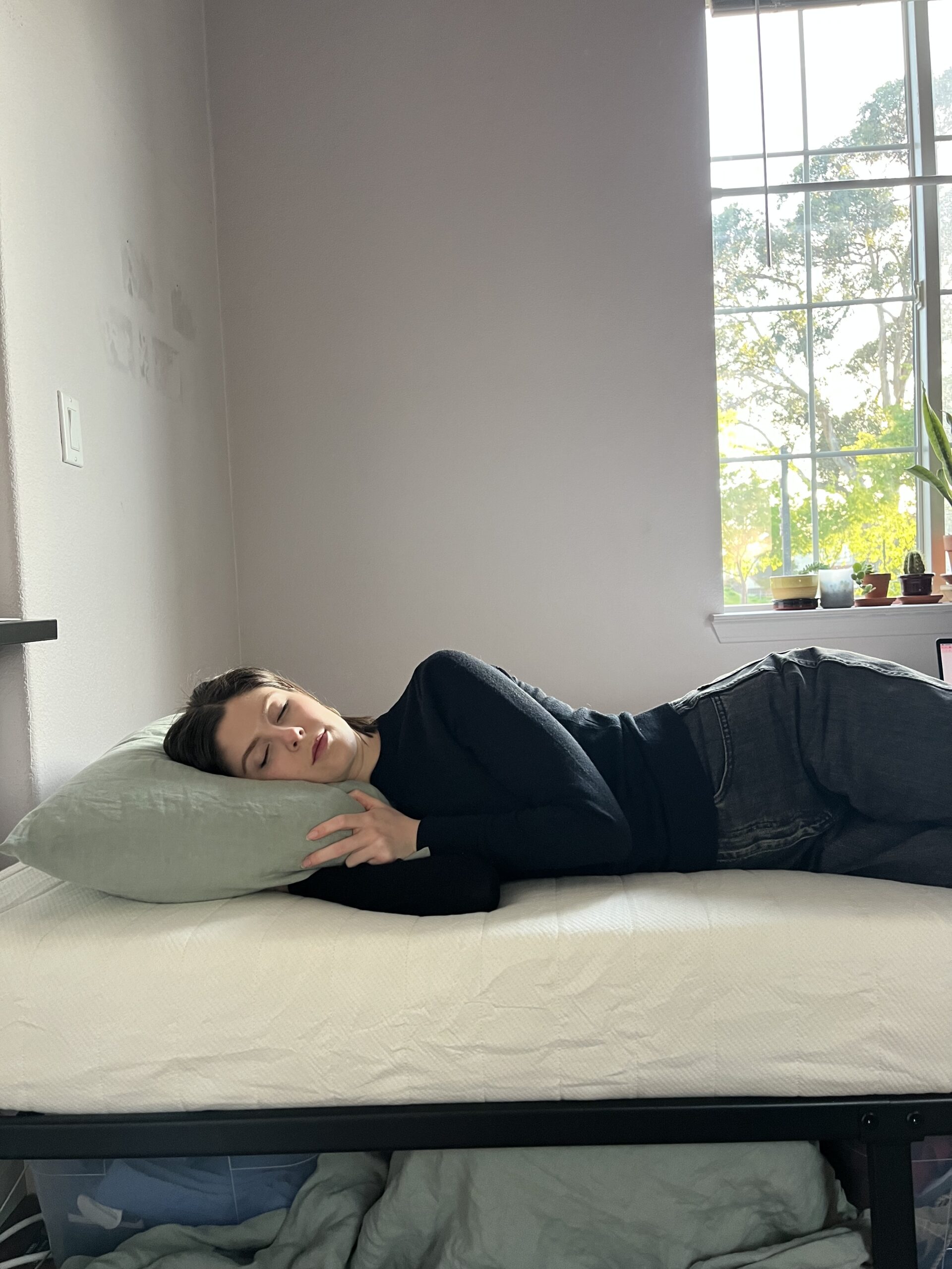 Person lying on their side, resting on a bed with a light green pillow. A window with potted plants is in the background, letting in natural light.