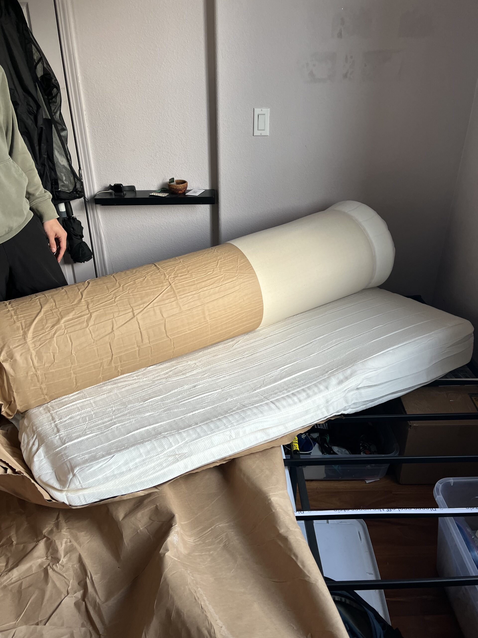 Unrolled mattress partially on a bed frame in a room, with a person standing nearby.