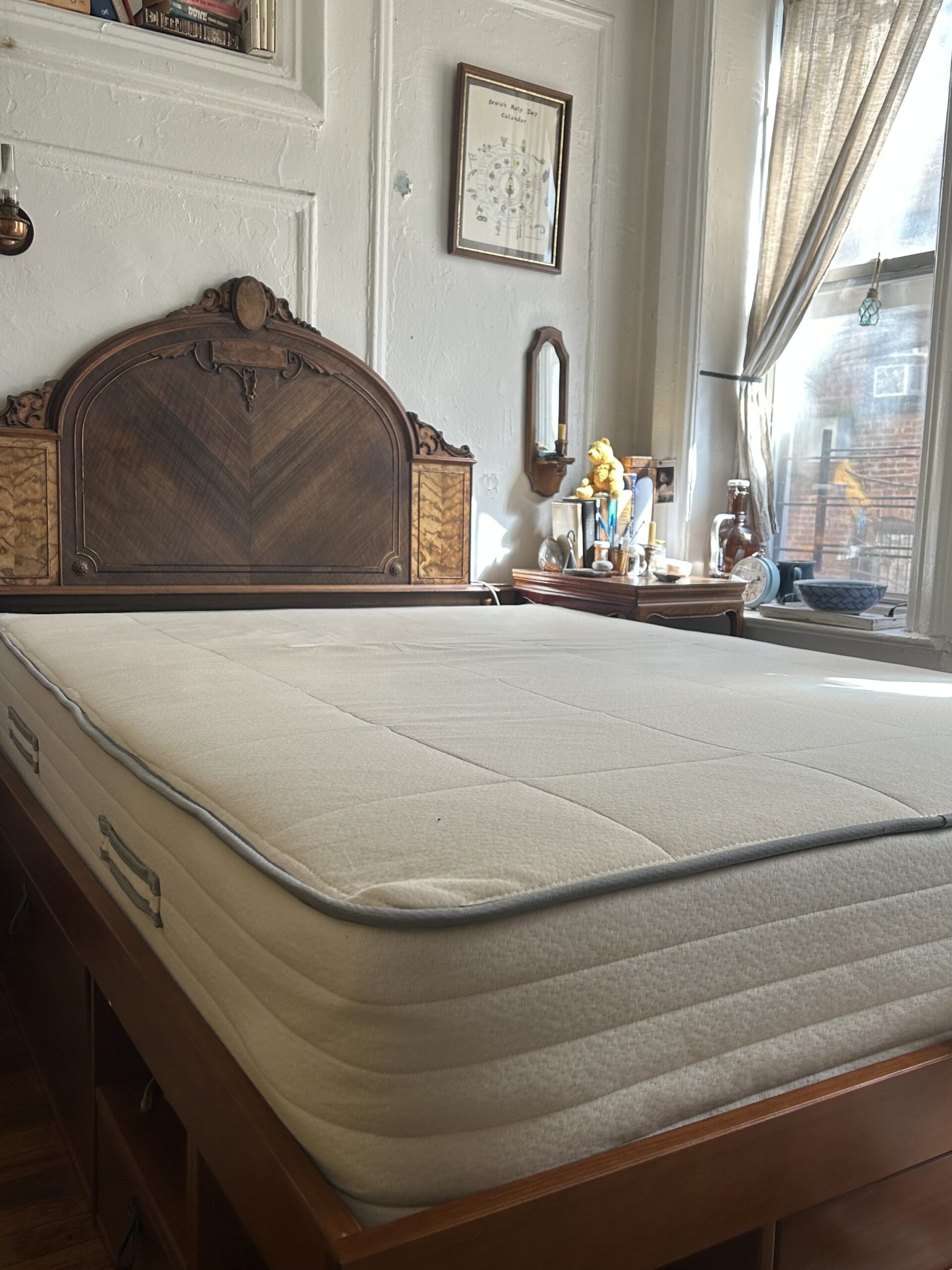 A neatly made bed with a thick mattress stands in a sunlit room with vintage decor, wooden headboard, and various items on the nightstand by a window.