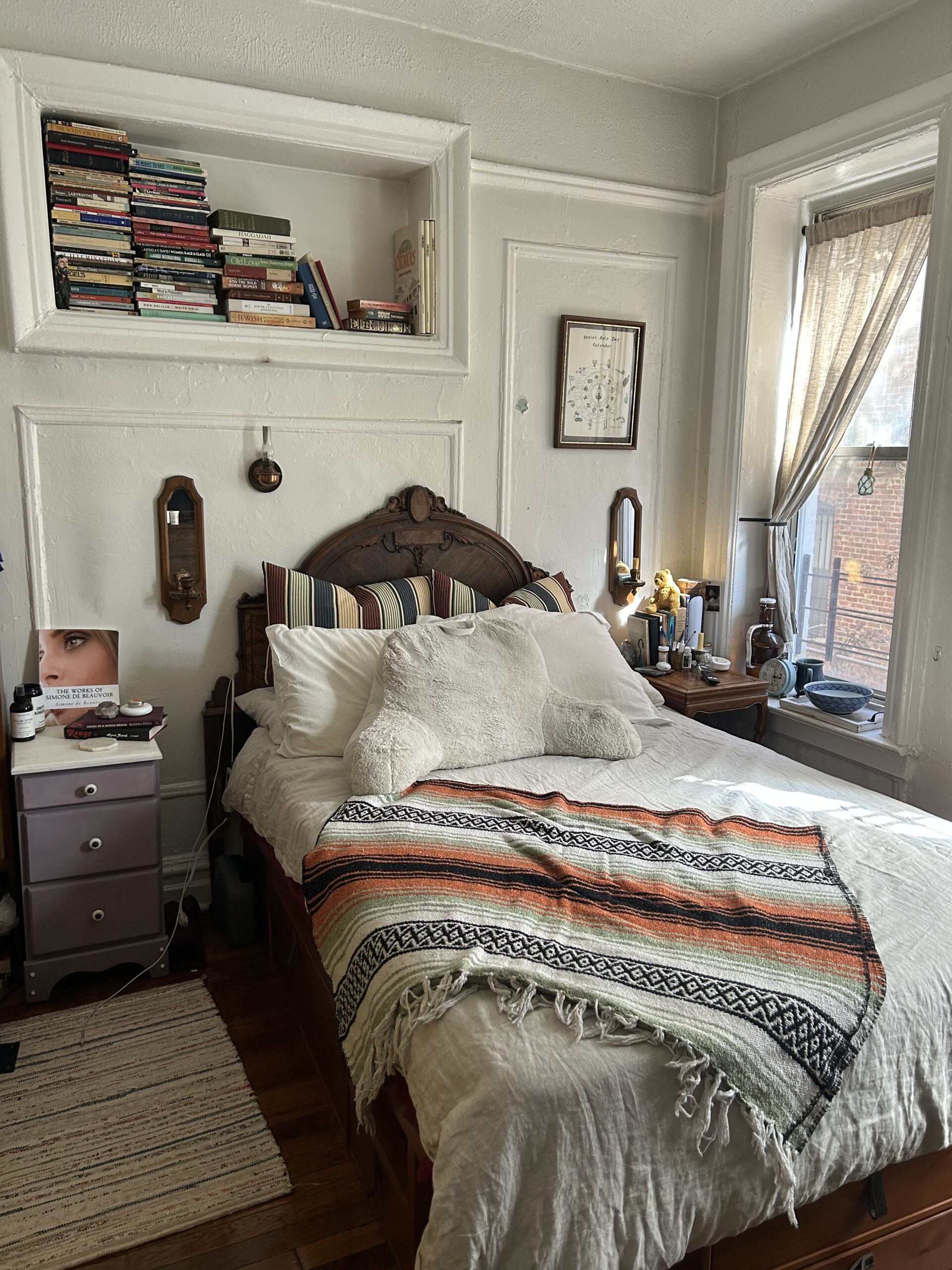 A neatly made bed with pillows and a striped throw blanket, flanked by nightstands and windows; shelves with stacked books are above the headboard.
