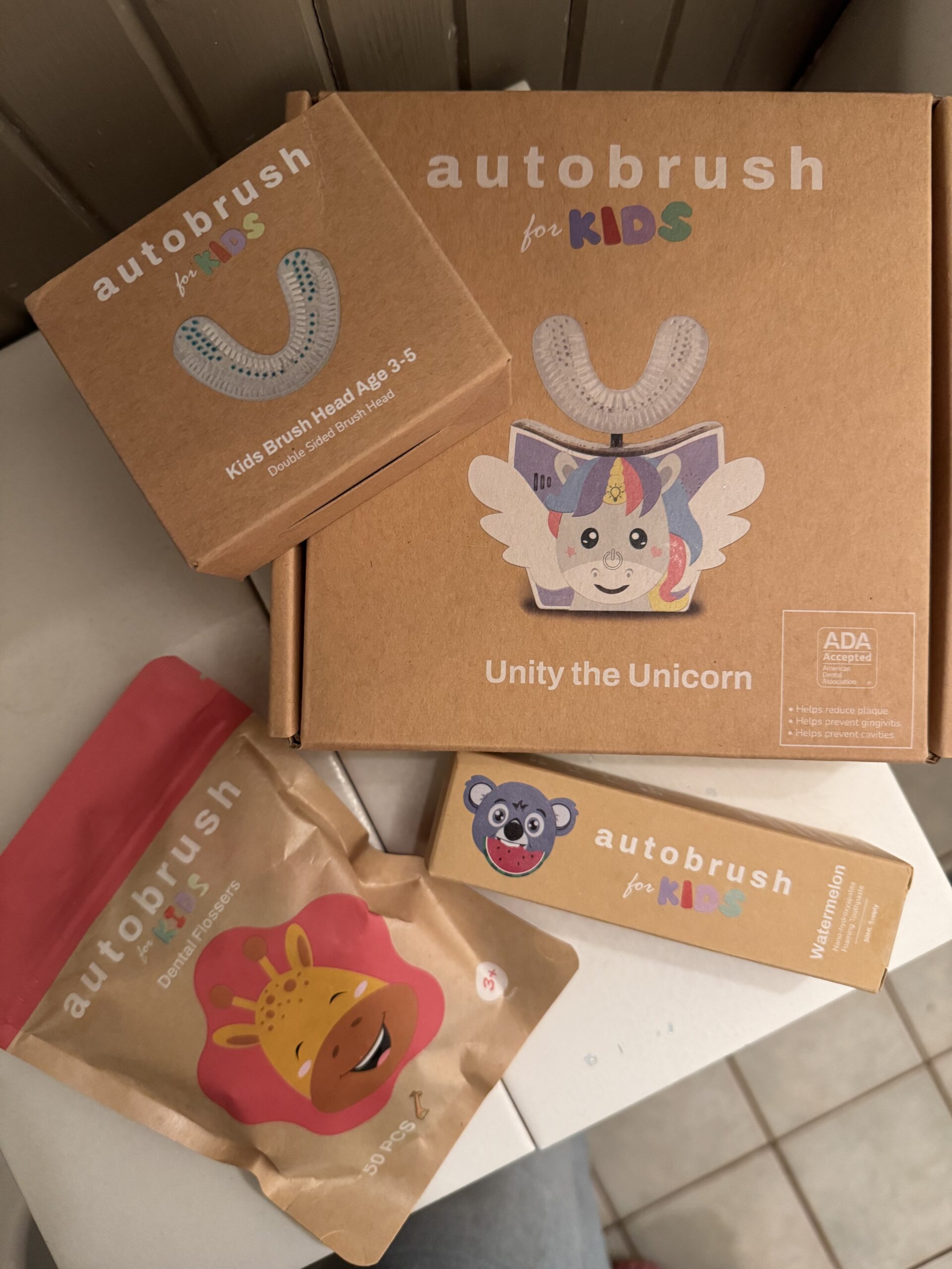 Boxes and pouches of children's automated toothbrush products featuring cartoon animals and a unicorn, labeled "autobrush for KIDS." The products are arranged on a surface.
