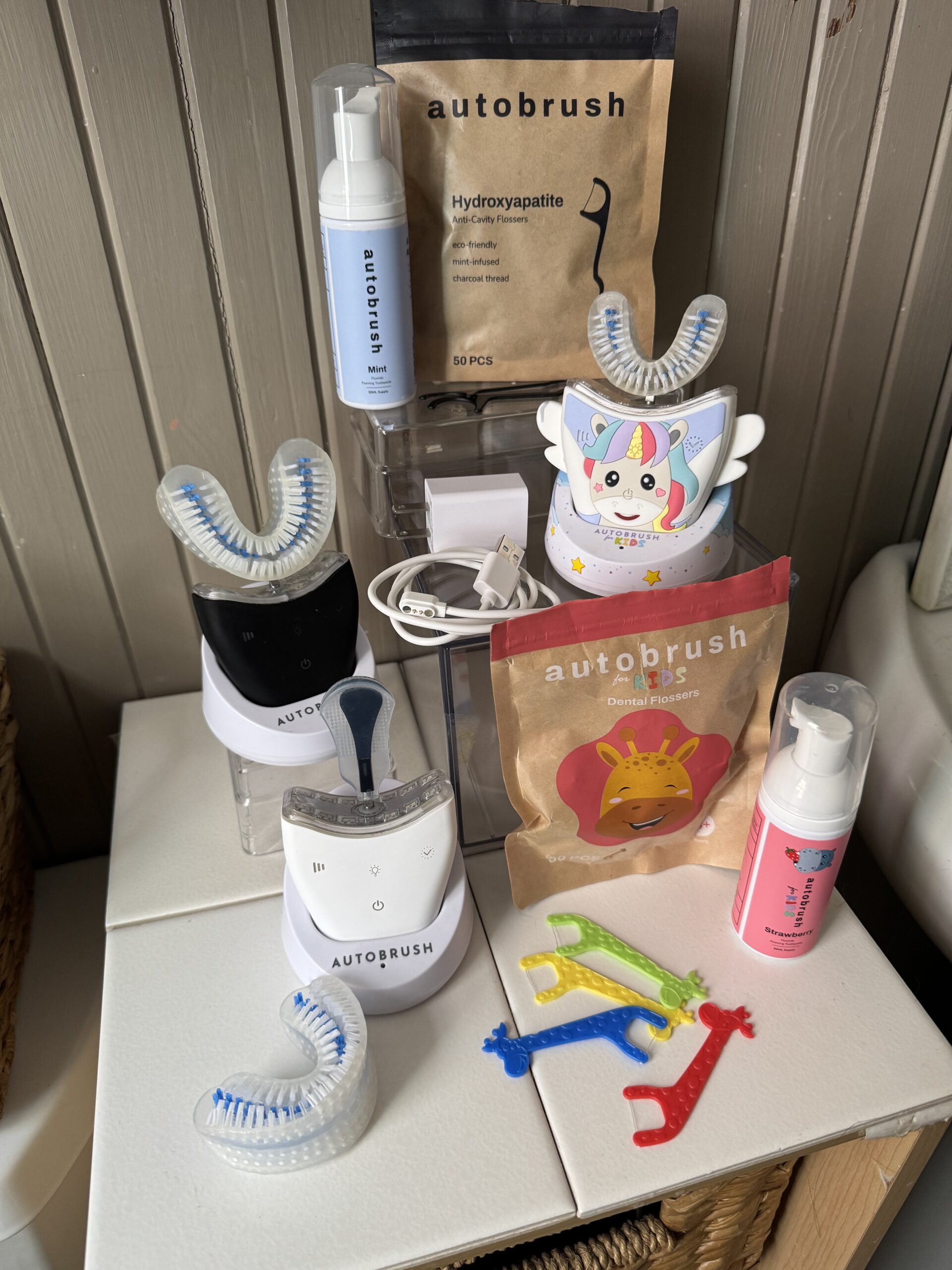 A collection of Autobrush products, including toothpaste powder, blue foam, various colorful toothbrushes, and two child-themed Autobrush devices on a wooden surface.