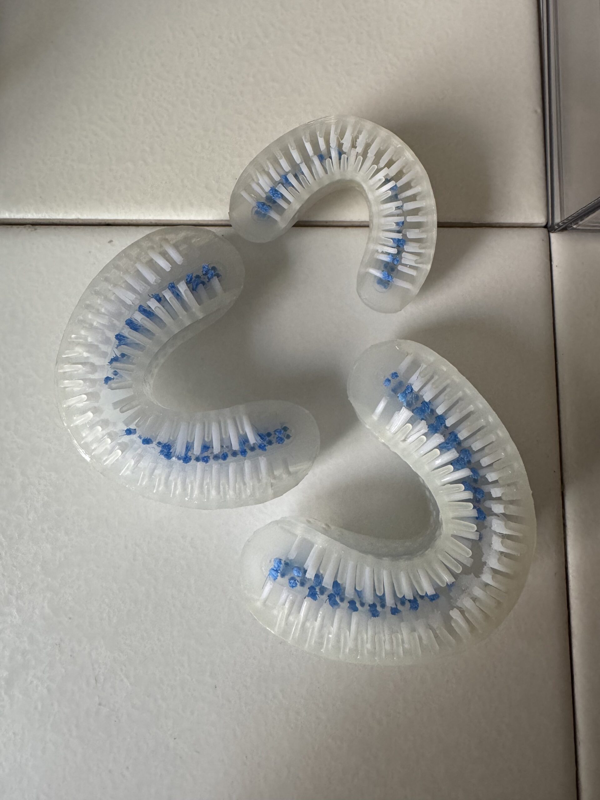 Three curved, clear plastic teeth-whitening mouthpieces with blue bristles are arranged on a white tiled surface.