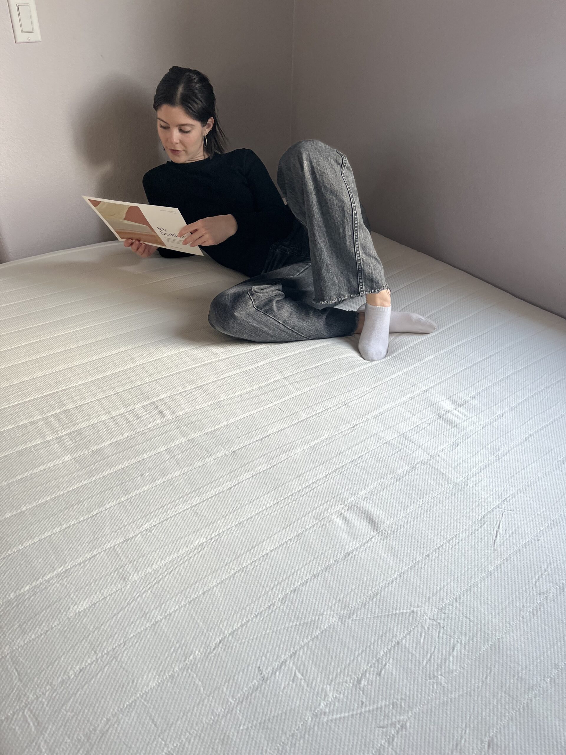 Person in casual attire reading a book while sitting on a large, unmade bed in a plain room.
