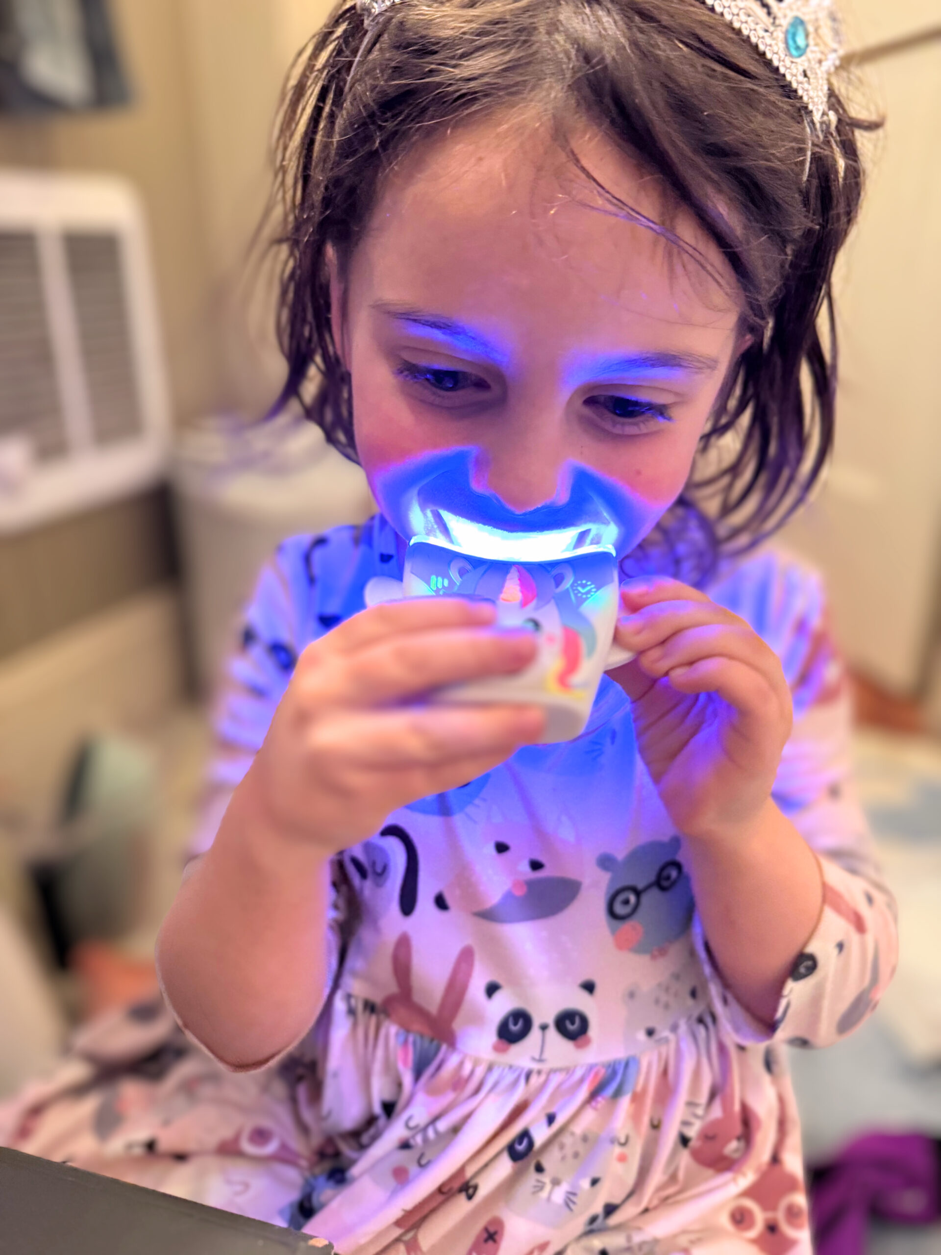 Child wearing a tiara looks at a glowing object held in both hands, illuminating their face with blue light. They are wearing pajamas with animal designs.