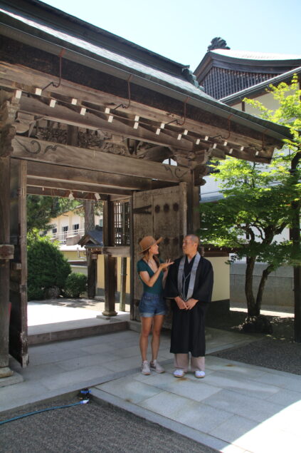 A woman in casual summer attire talks to a man wearing traditional black robes under a wooden temple gate with intricate carvings. A tree and building are visible in the background.