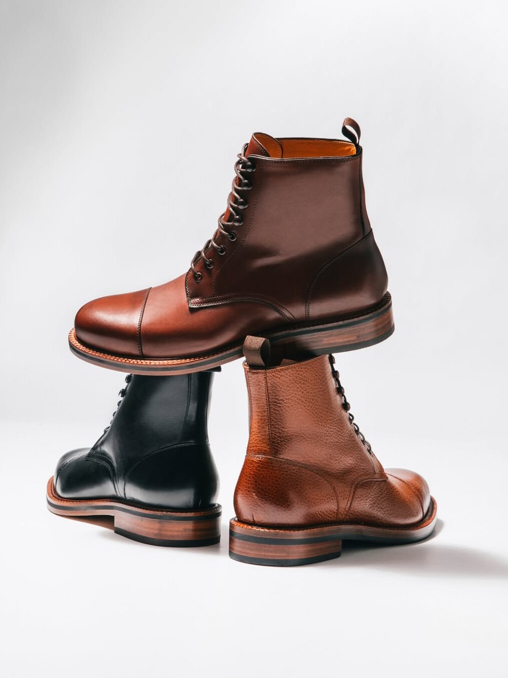 Three leather lace-up boots—two brown and one black—are stacked in a balanced arrangement against a plain white background.