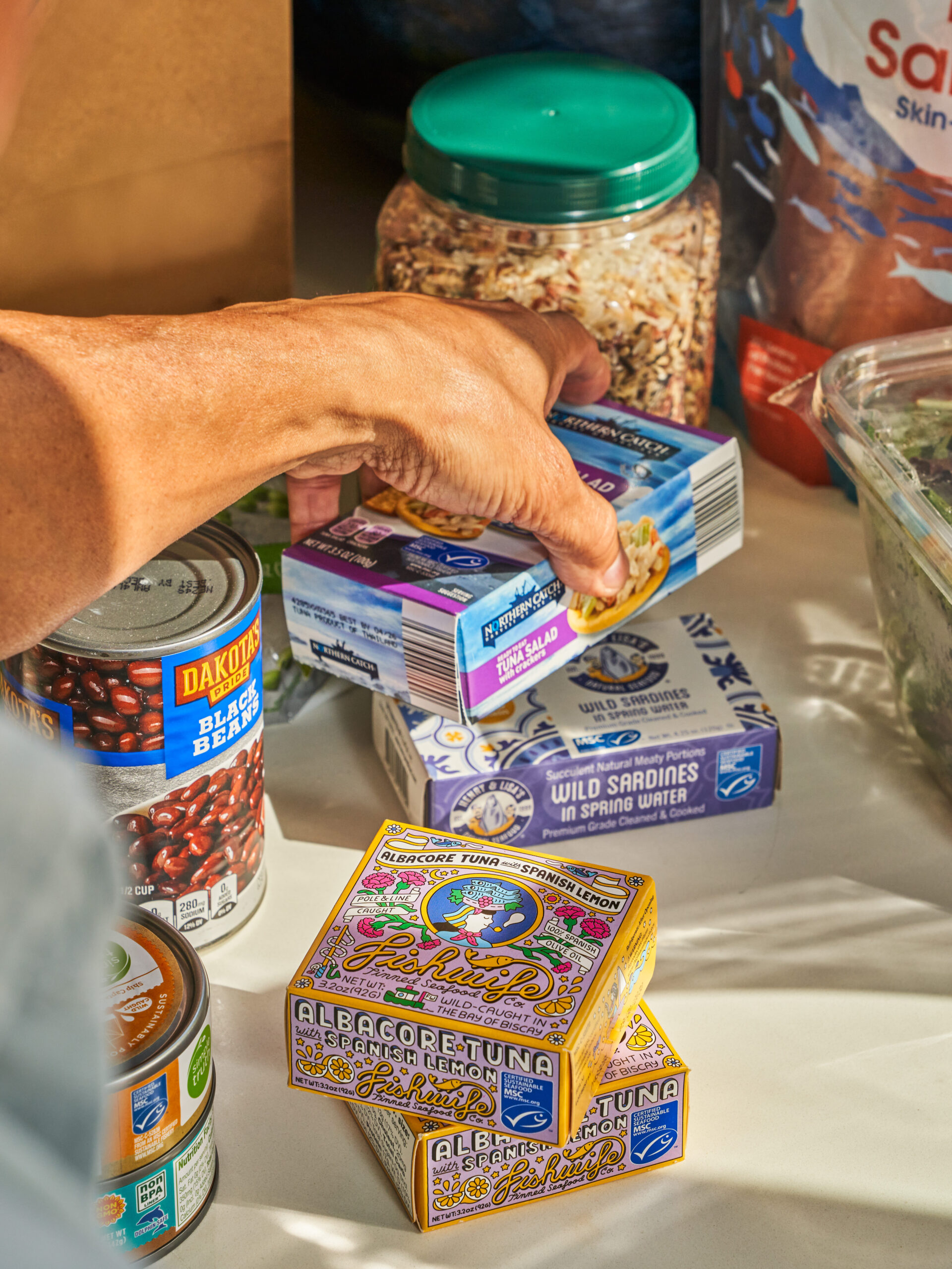 A person selects a package of wild sardines from a variety of canned goods, salad greens, dry oats, and a bag of pink salmon fillets on a kitchen counter.
