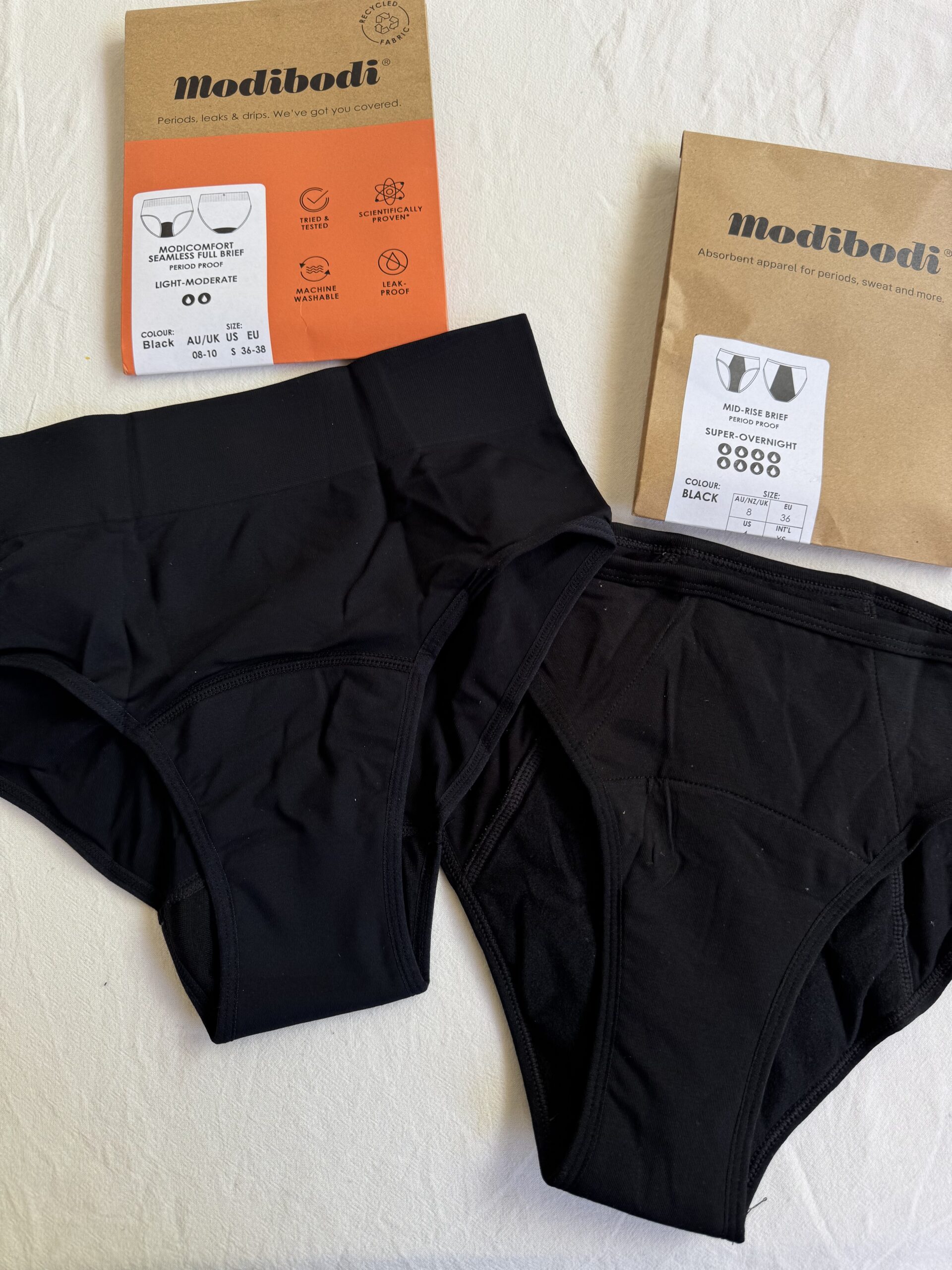 Two pairs of black Modibodi underwear on a white surface, with packaging showing product details and features.