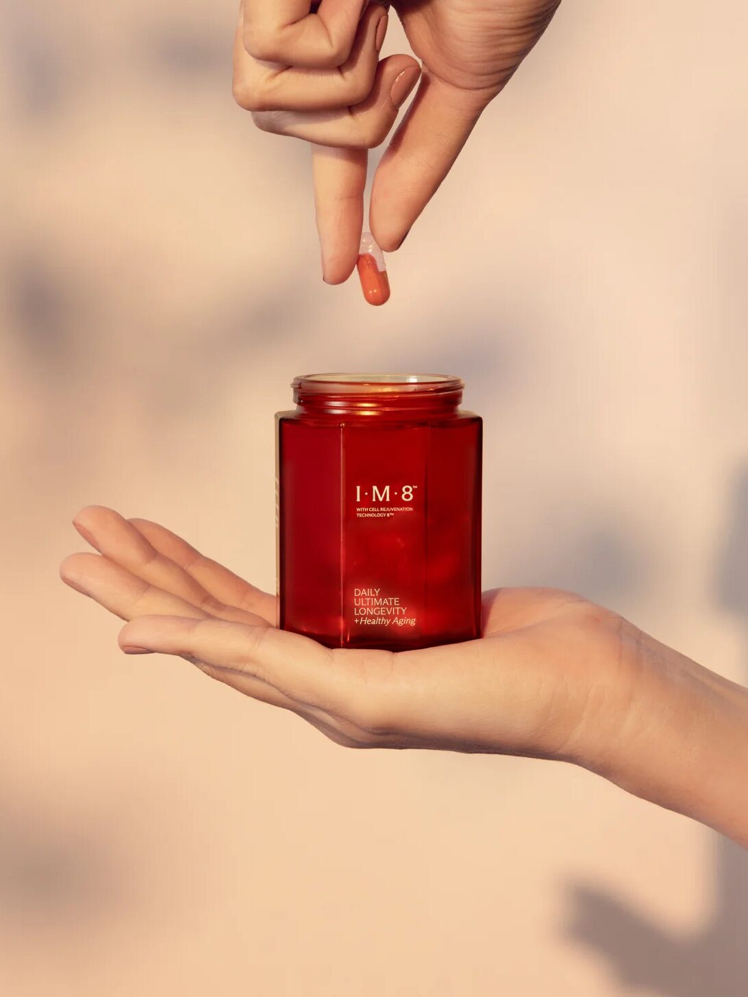 A hand holds a red jar while another hand hovers above, releasing a small capsule into the jar. The background is neutral and out of focus.