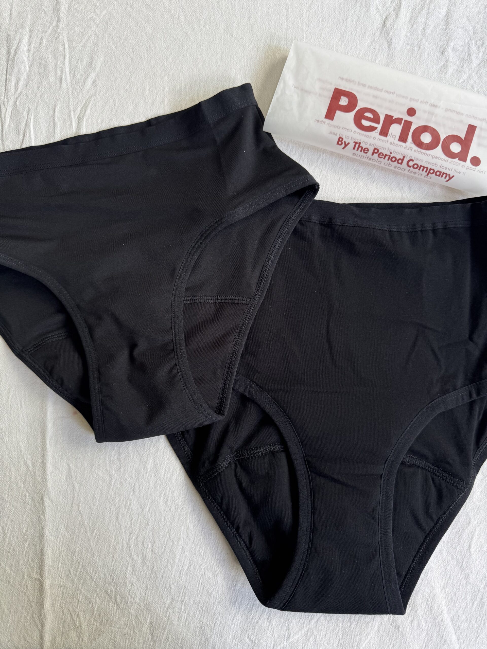 Two black underwear pieces are laid out on a white surface next to a branded envelope labeled "Period. By The Period Company.