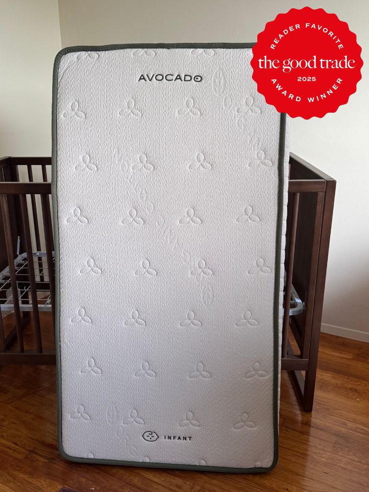 A crib mattress labeled "Avocado Infant" stands upright in a room with wooden furniture; a 2025 "The Good Trade" award badge is displayed in the top right corner.
