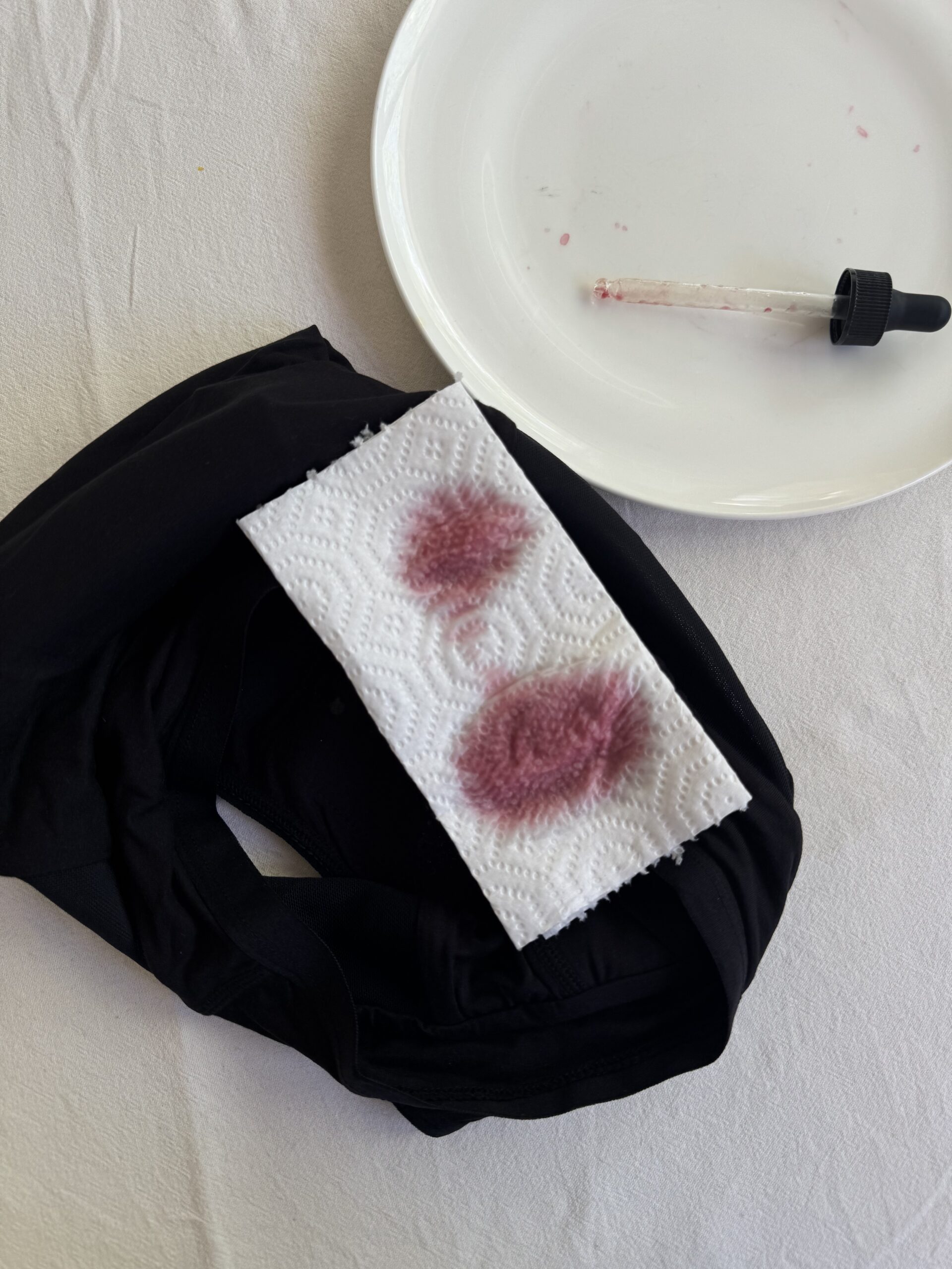 A fabric with two reddish stains on a white paper towel, next to a dropper and an empty white plate, placed on a white surface.