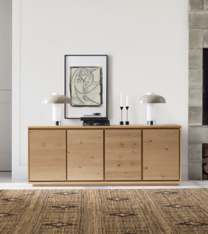 A wooden sideboard with two lamps, stacked books, and two candlesticks stands against a white wall with framed abstract art above, next to a stone fireplace.