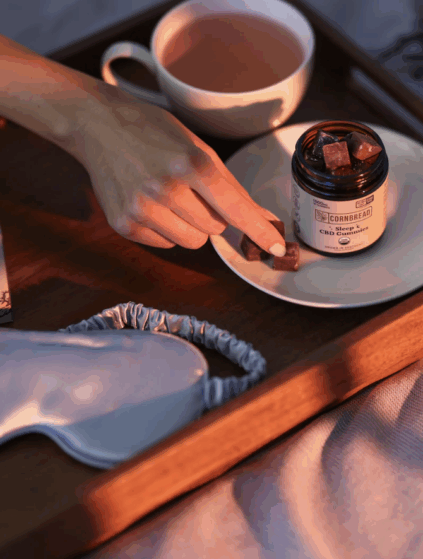 A hand picks a gummy from a plate beside a jar labeled "Cornbread CBD Gummies," with a cup of tea and a sleep mask on a wooden tray.
