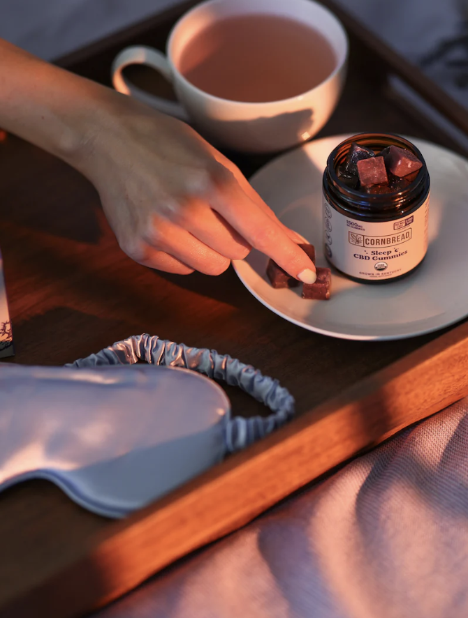 A hand picks a gummy from a plate beside a jar labeled "Cornbread CBD Gummies," with a cup of tea and a sleep mask on a wooden tray.