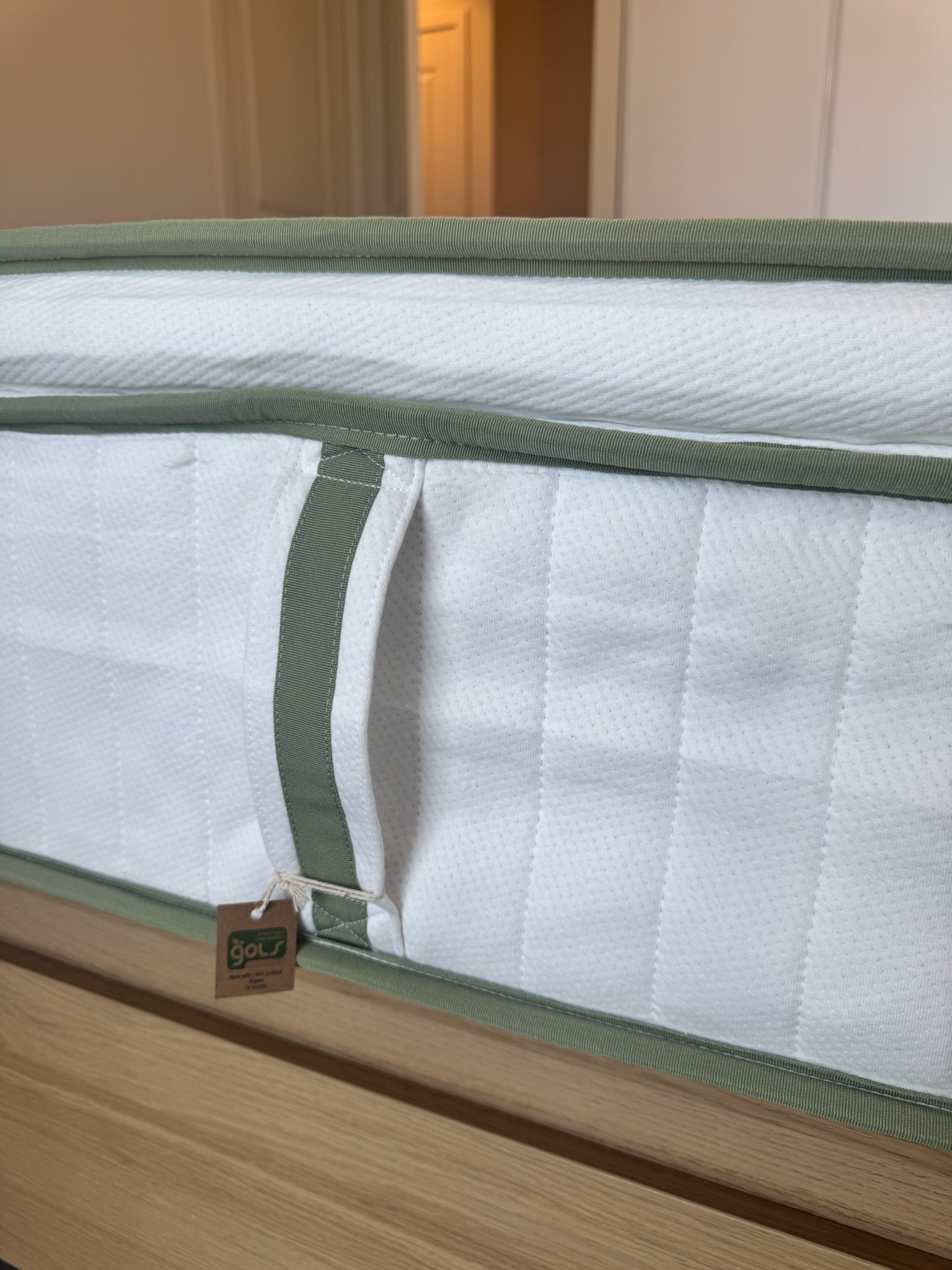 Close-up of a mattress with a green trim, a fabric handle, and a small brown tag labeled "golu," resting on a light wooden surface in a bedroom.