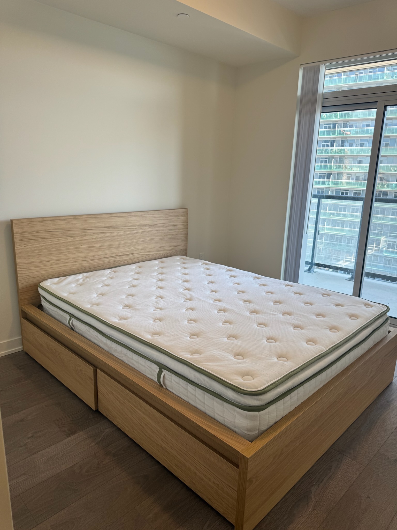 A wooden bed frame with storage drawers under a mattress in a minimally furnished room with a large window and cityscape view.
