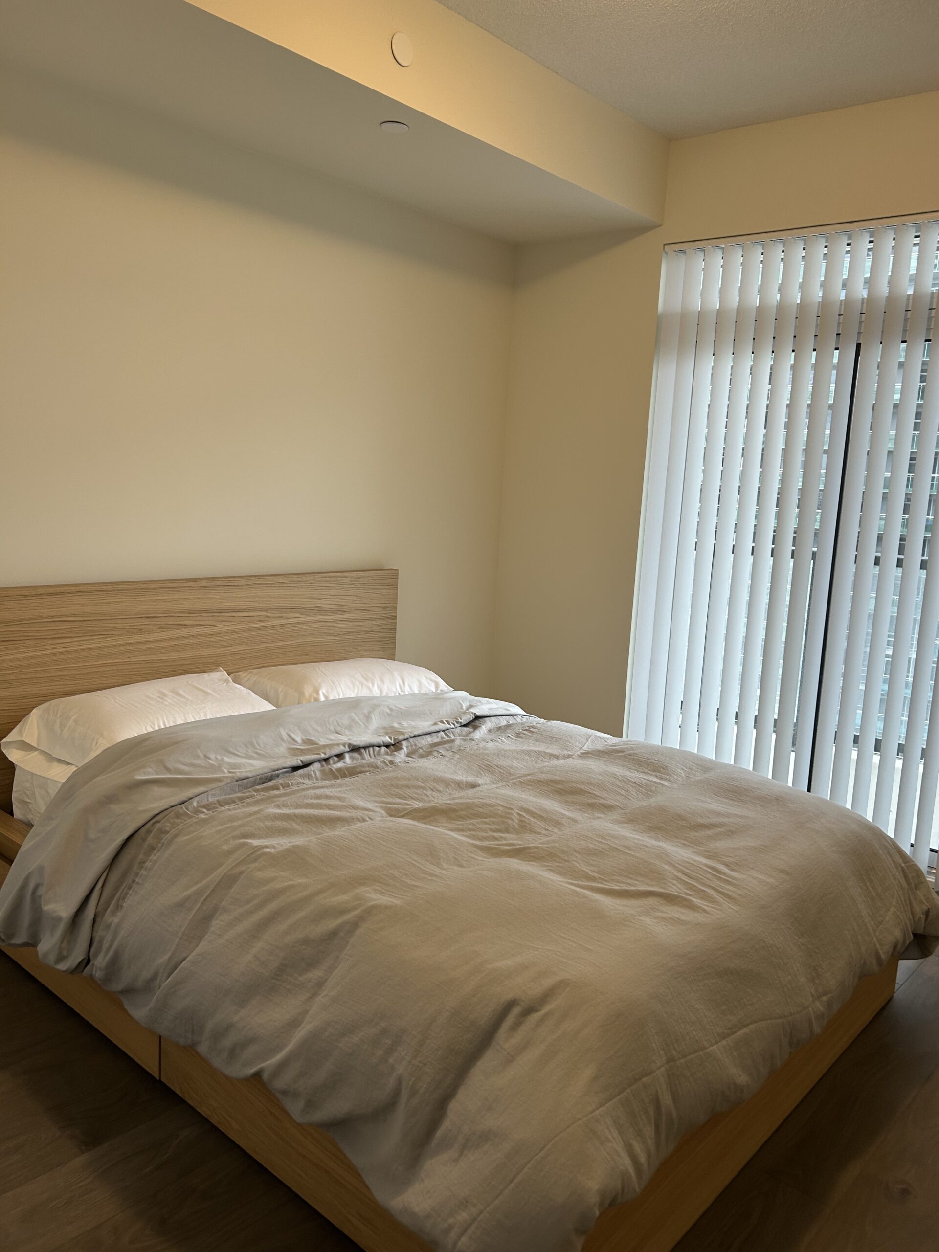A neatly made bed with light gray bedding and two pillows is positioned beside a window with vertical blinds in a simple, modern bedroom.