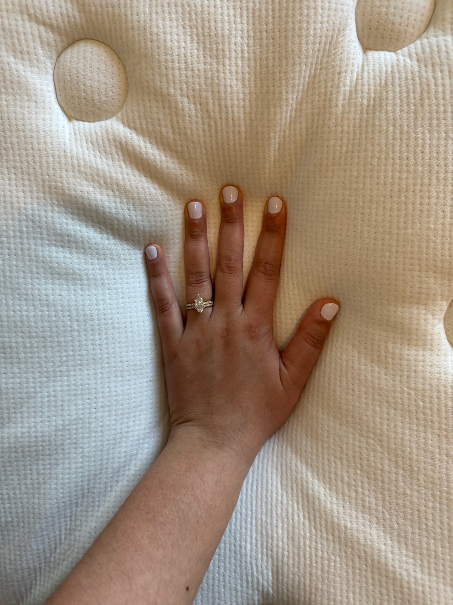 A hand with white-painted nails and a diamond ring presses into a textured, white mattress with visible indents.