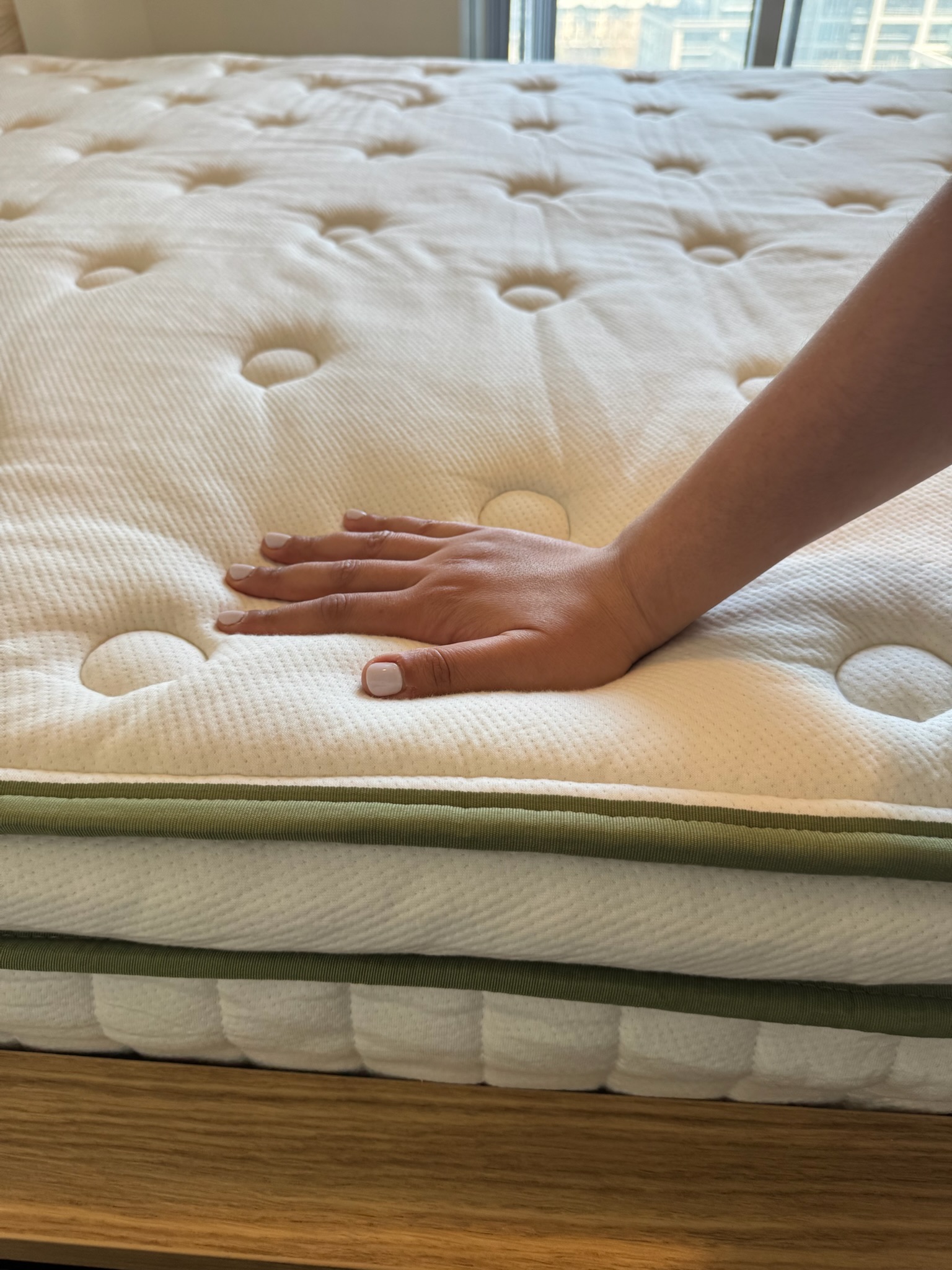 A hand pressing down on a white, tufted mattress with green trim, showing the mattress’s softness and texture.