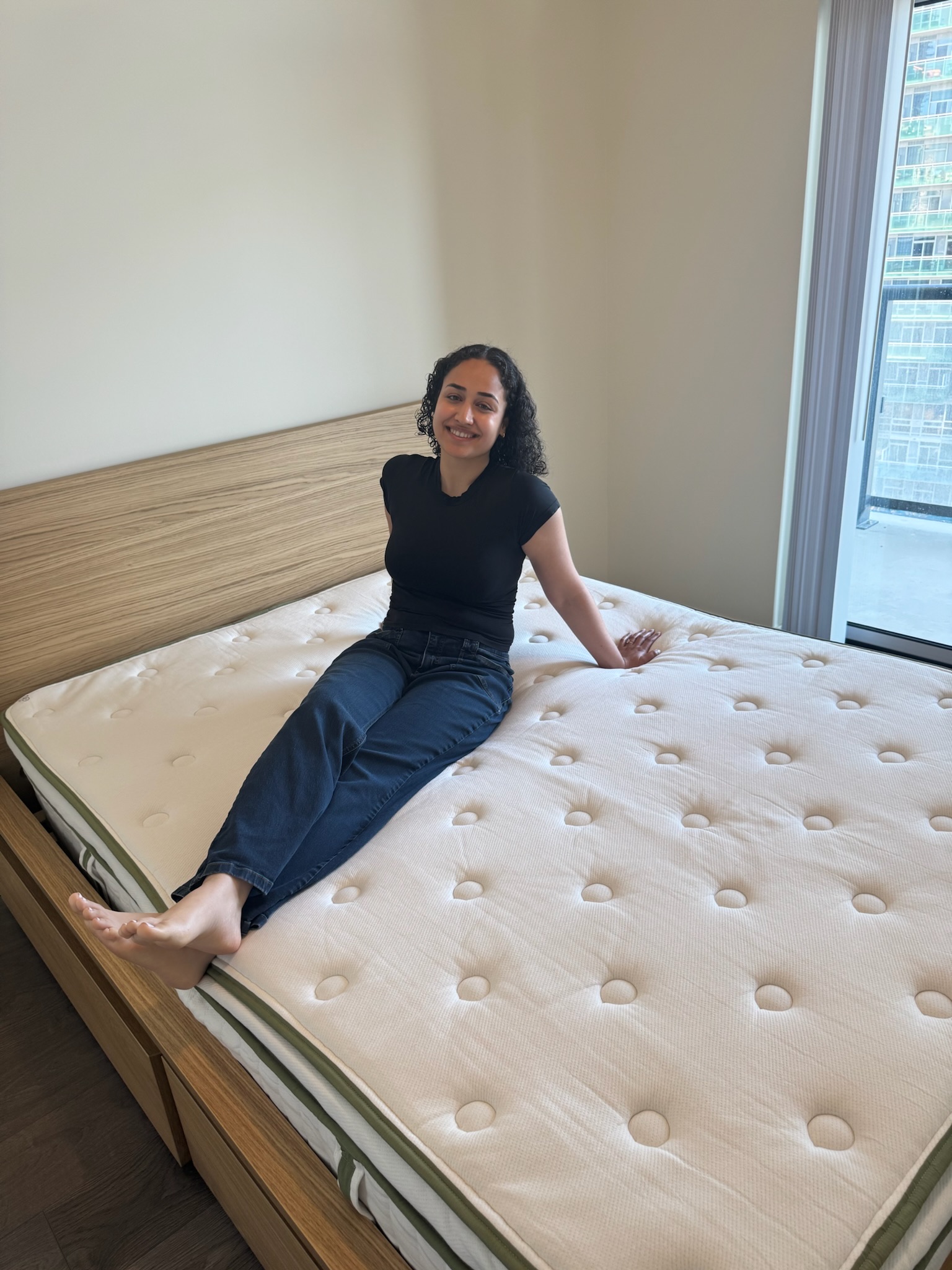 A person sits on a neatly made bed with a plain mattress in a modern, bright bedroom with large windows.