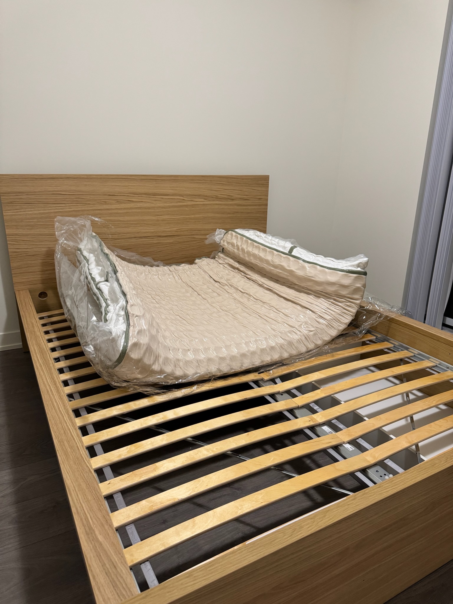 A deflated mattress in plastic packaging is placed on top of a wooden slatted bed frame in a minimally furnished room.