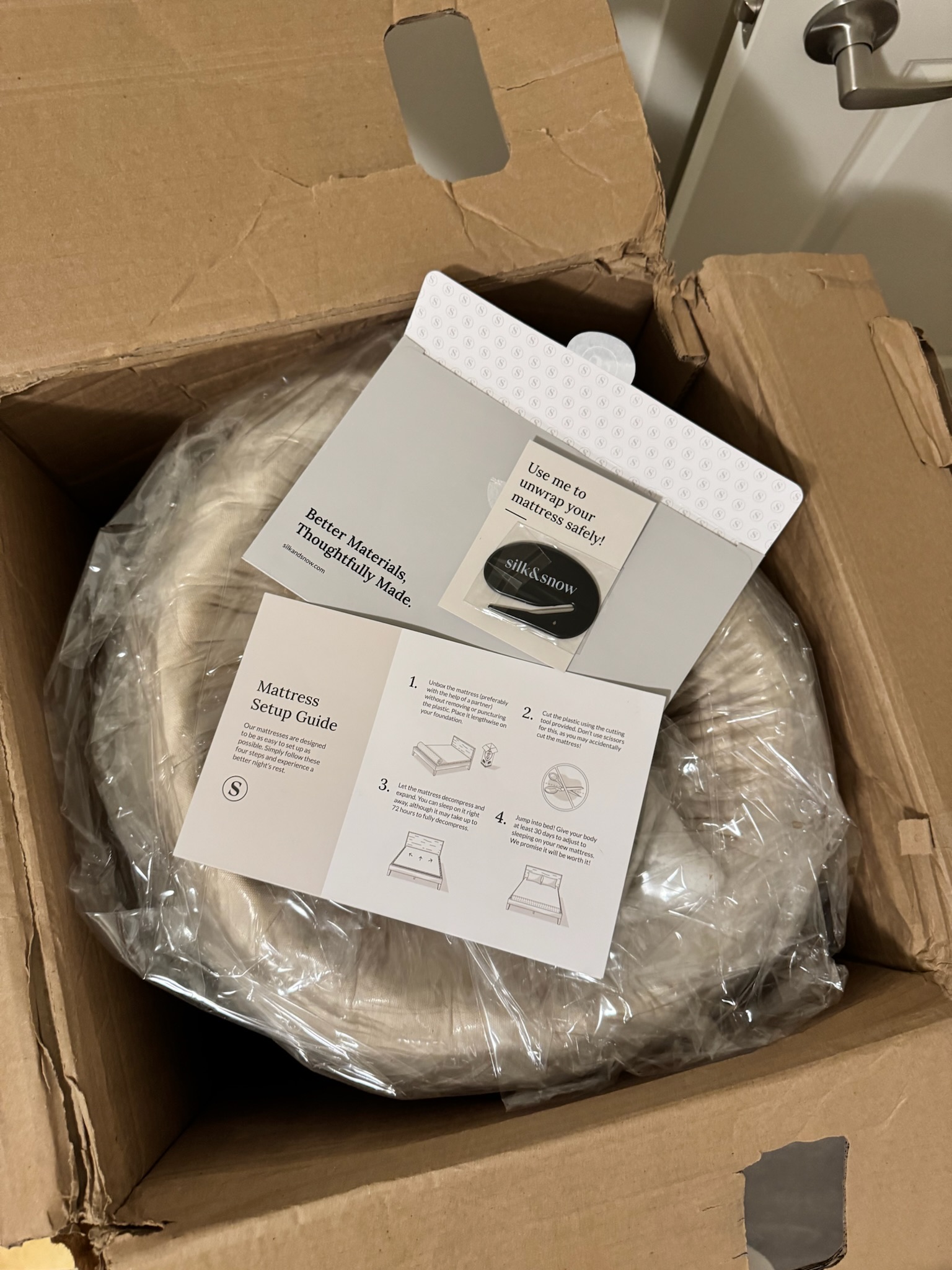 A rolled-up mattress in plastic packaging inside an open cardboard box, with a mattress setup guide and informational pamphlet placed on top.