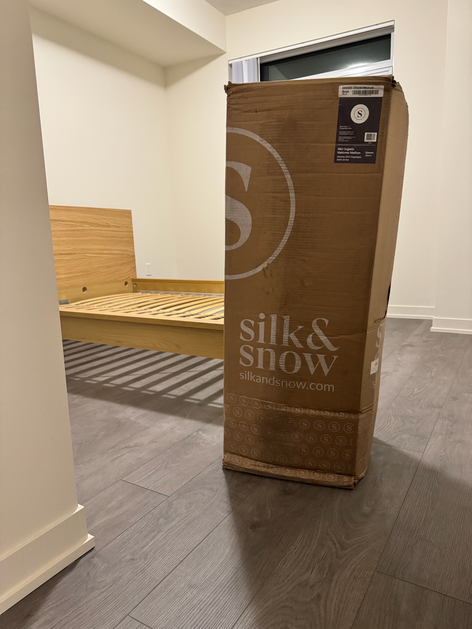 A boxed Silk & Snow mattress stands upright on a wooden floor next to an unassembled bed frame in a minimally furnished room.