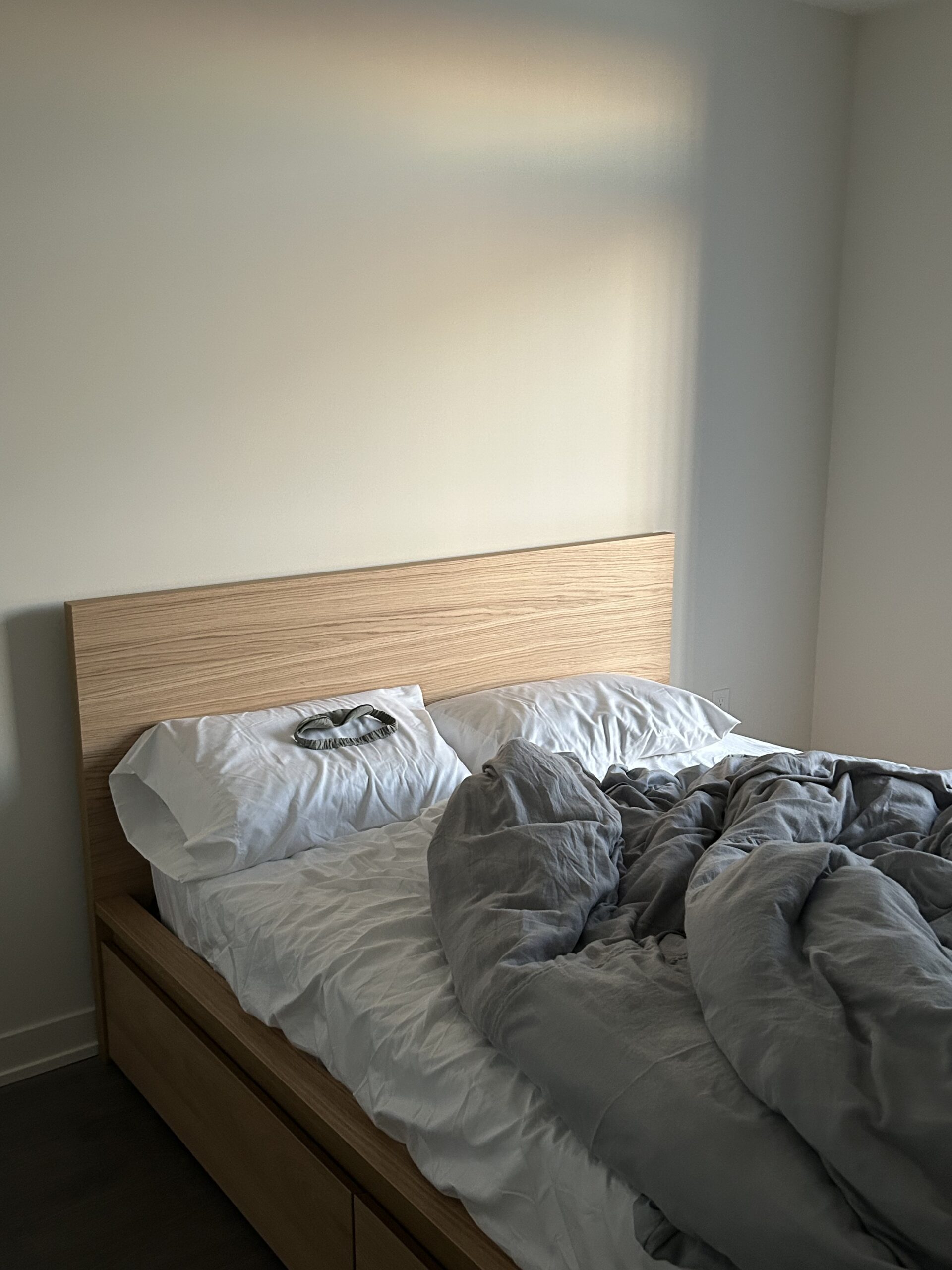 A modern bed with a wooden headboard, white sheets, and a gray comforter partially pulled back in a bright, minimally furnished room.