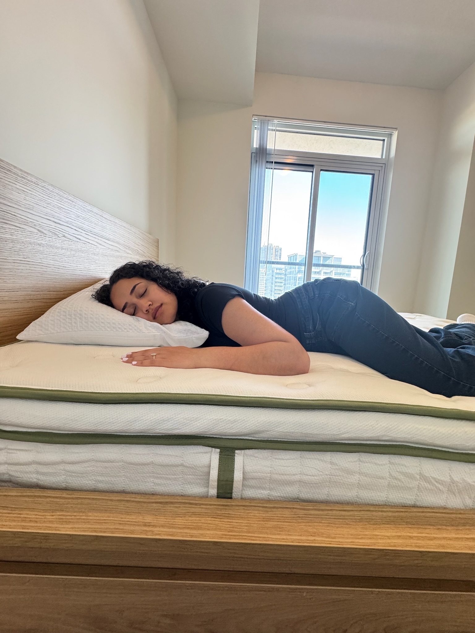 A woman lies on her stomach on a bed with a white mattress and pillow, facing a window with a city view in the background.