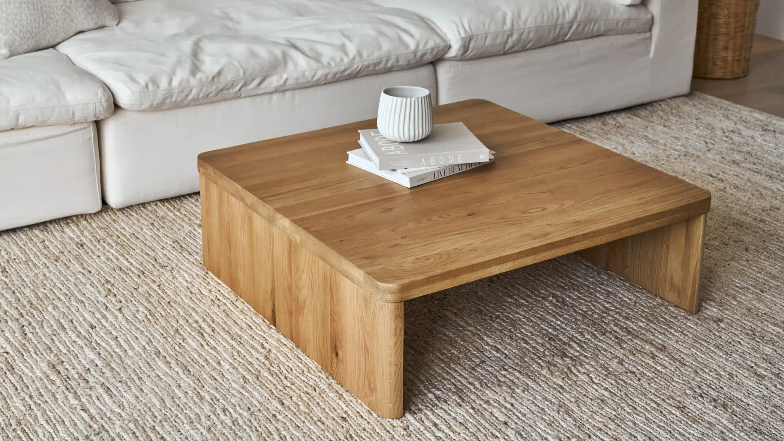 A wooden coffee table with a stack of two books and a textured white cup sits on a light woven rug in front of a white sofa.