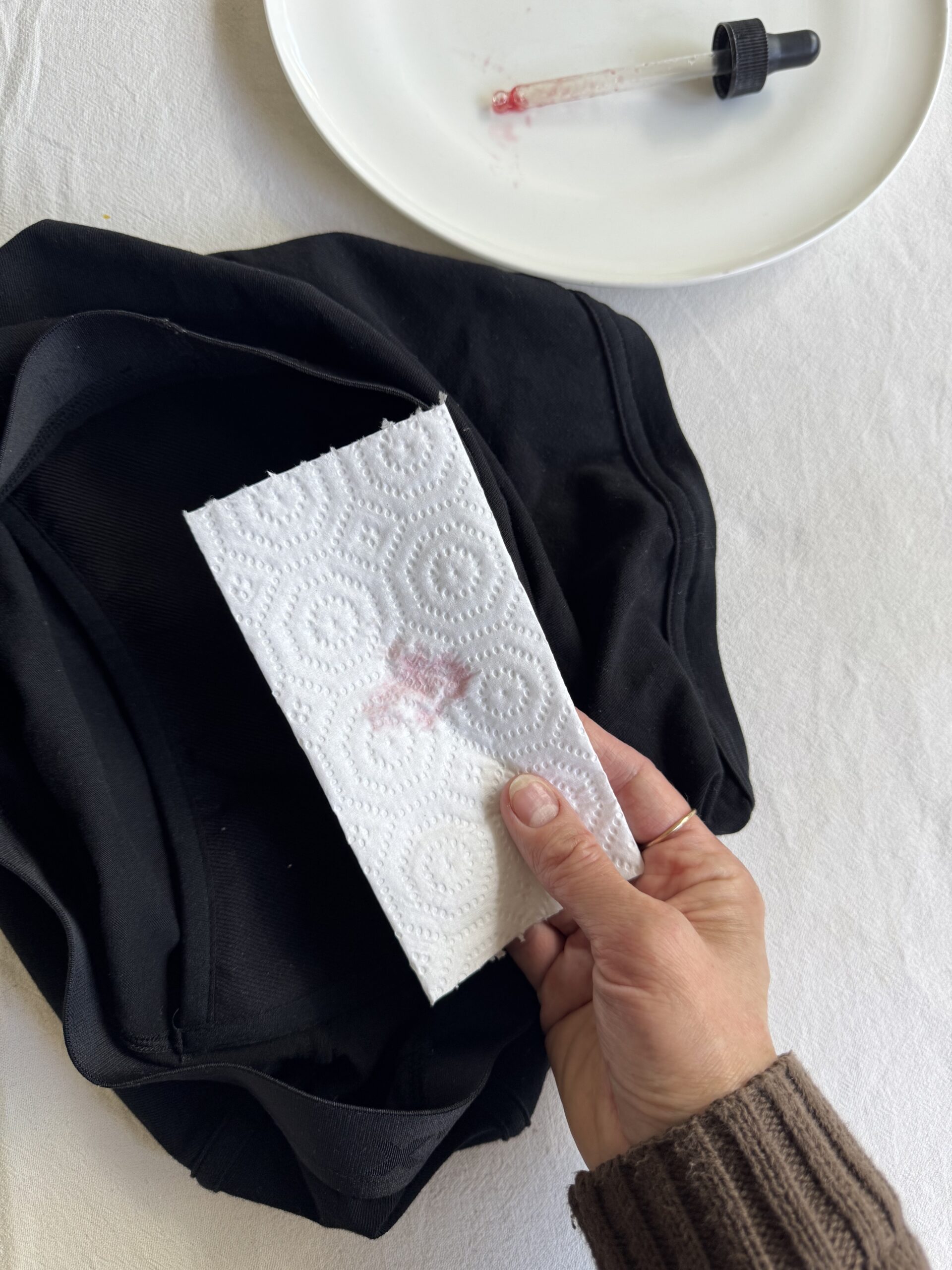 A hand holds a paper towel with a red stain over a black fabric item. A white plate and a dropper are visible in the background.