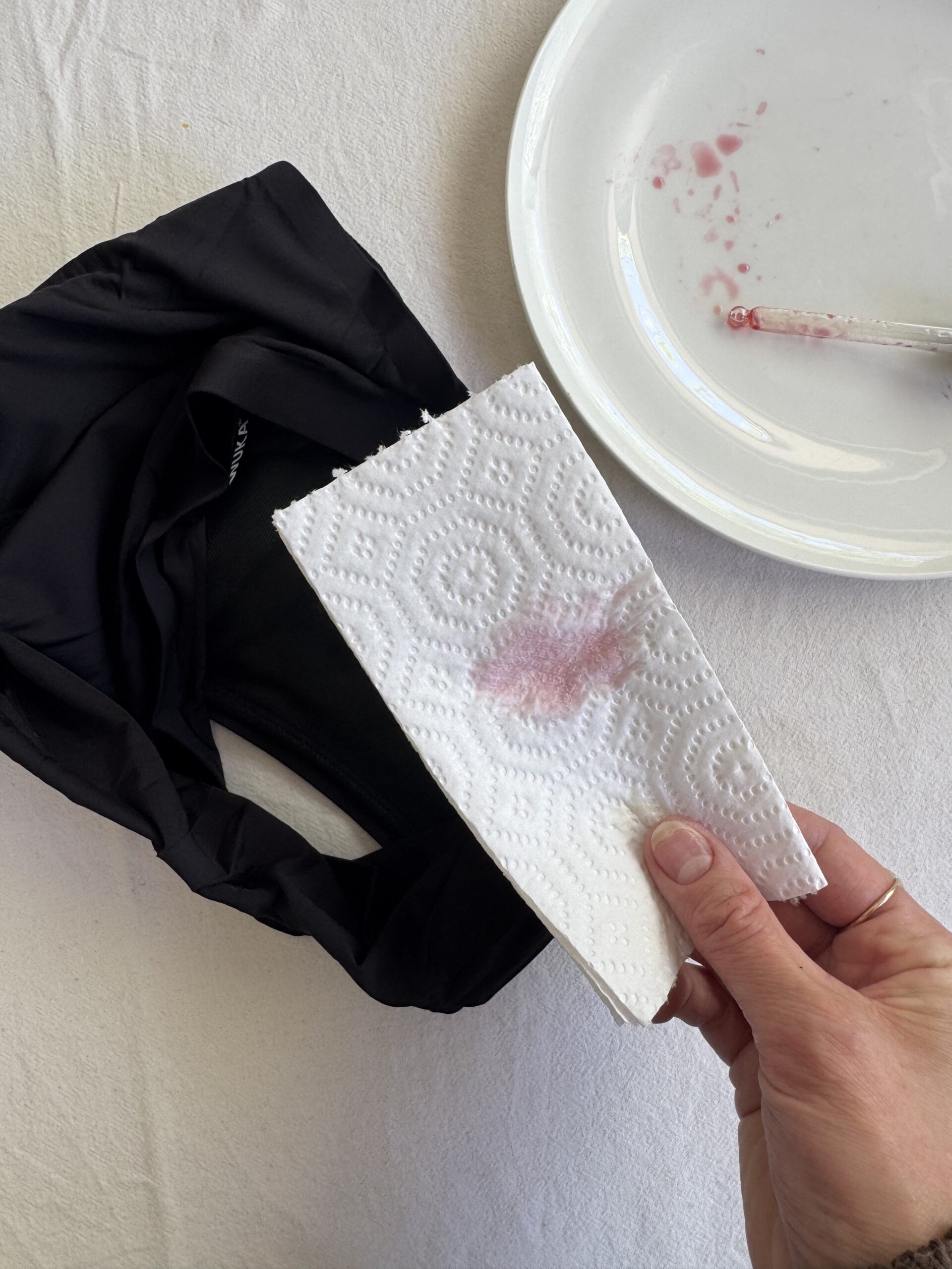 Hand holding a paper towel with a pink stain, next to a black fabric item, a white plate, and a glass tube containing pink residue on a white surface.