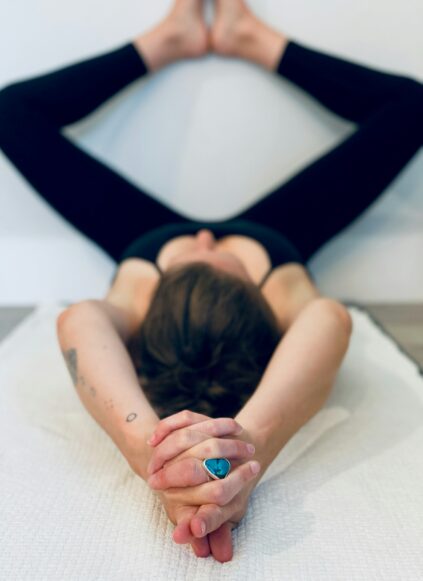 Person lying on their back on a mat with legs spread against a wall and arms extended overhead, hands clasped, wearing black leggings and a blue ring.