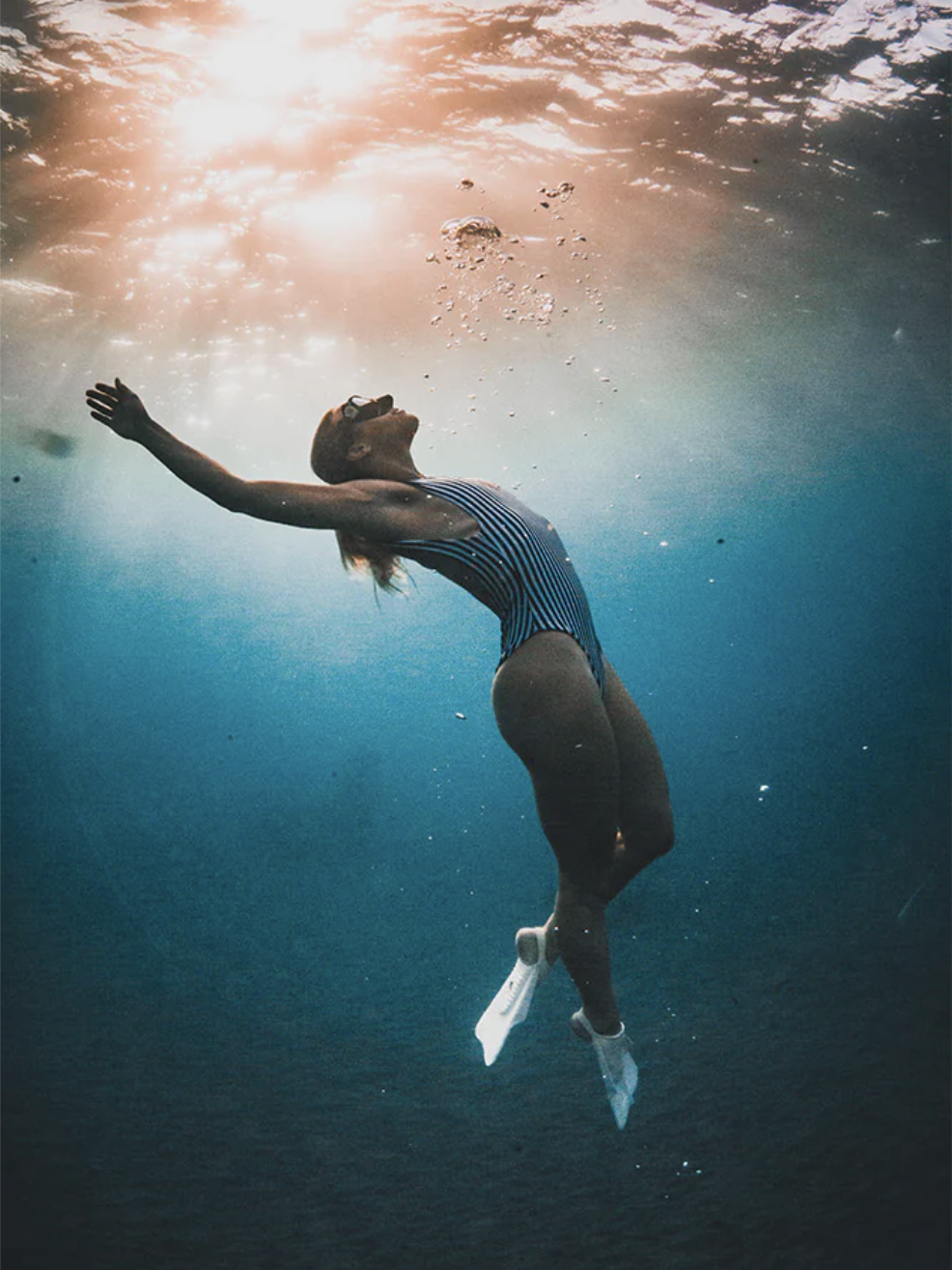 A person in a striped swimsuit and fins swims underwater, arms outstretched, with sunlight filtering through the water above.